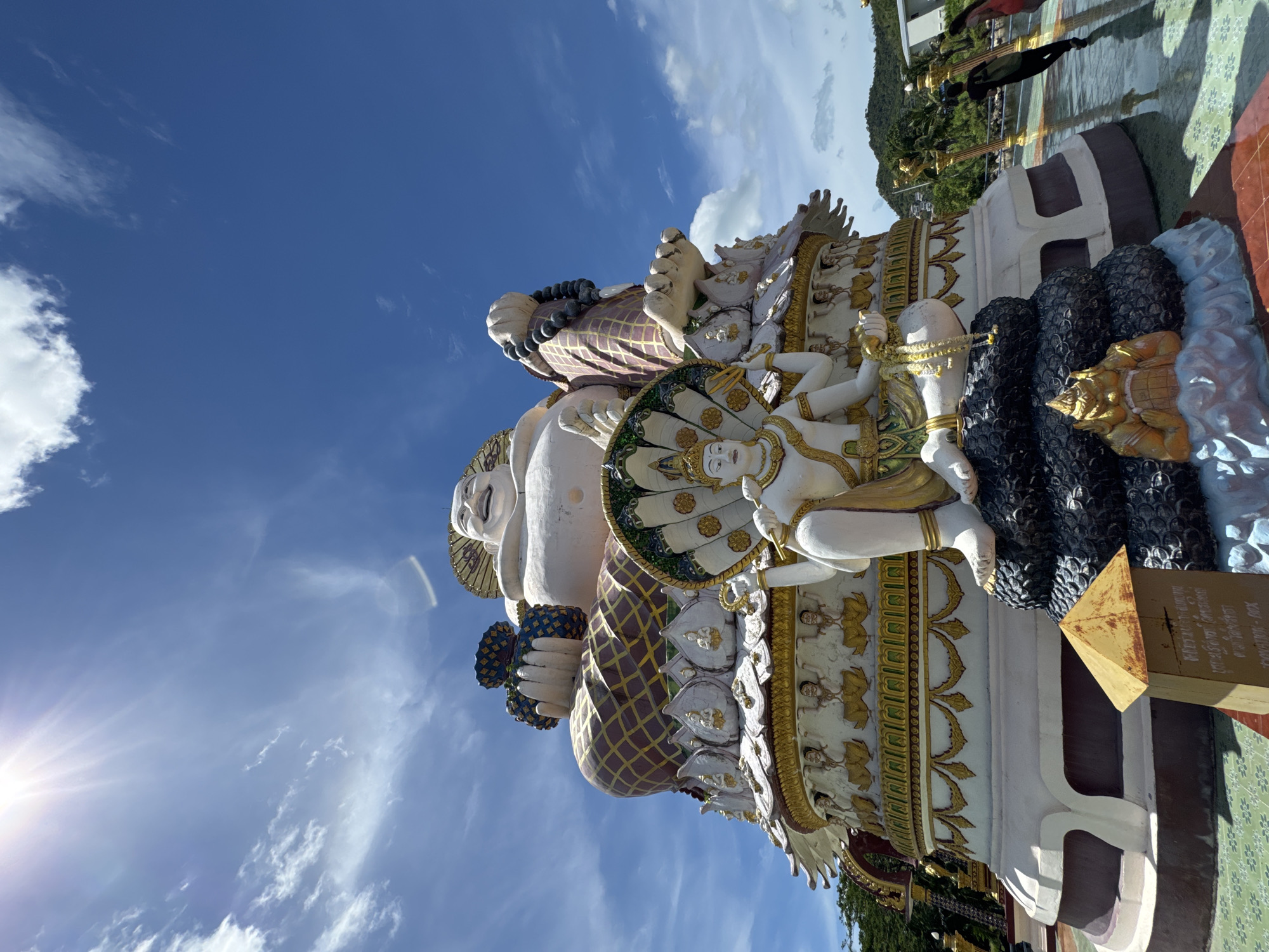 The image shows a large, ornate statue of a smiling Buddha or deity in Baan Plai Laem, Thailand, under a bright blue sky with scattered clouds. The statue is multi-tiered, with a large, rotund figure at the top holding a bag of treasures. Below this figure is another seated deity with multiple arms and a serpent-like headdress. The lower levels of the statue are decorated with smaller figures and intricate golden detailing. In the foreground, there is a small, weathered wooden box with inscriptions. To the right of the statue, two individuals are walking away from the camera on a patterned tiled surface. In the background, lush green hills and some buildings are visible. The sun is shining brightly in the upper left corner, creating lens flare.