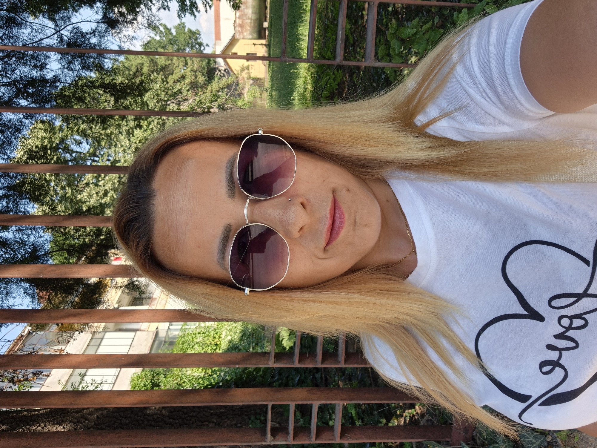 A blonde woman with sunglasses and a white t-shirt with a heart design stands in front of a metal fence. The background shows a building and trees.