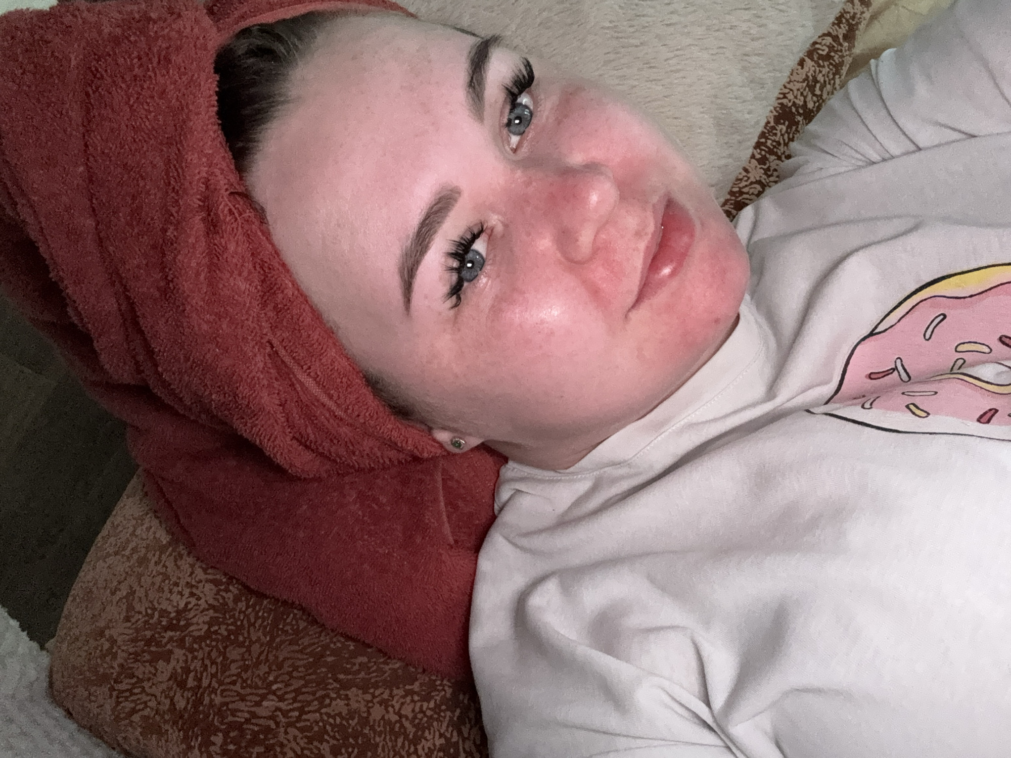 A woman with fair skin and blue eyes is lying on a patterned surface, possibly a bed or a cushion. She has a red towel wrapped around her head. Her face is flushed, particularly her cheeks and nose. She wears eyelash extensions and has well-defined eyebrows. She is wearing a white t-shirt with a pink donut print on the upper right side. The location cannot be confirmed as being Ol'khovochka, Russia.