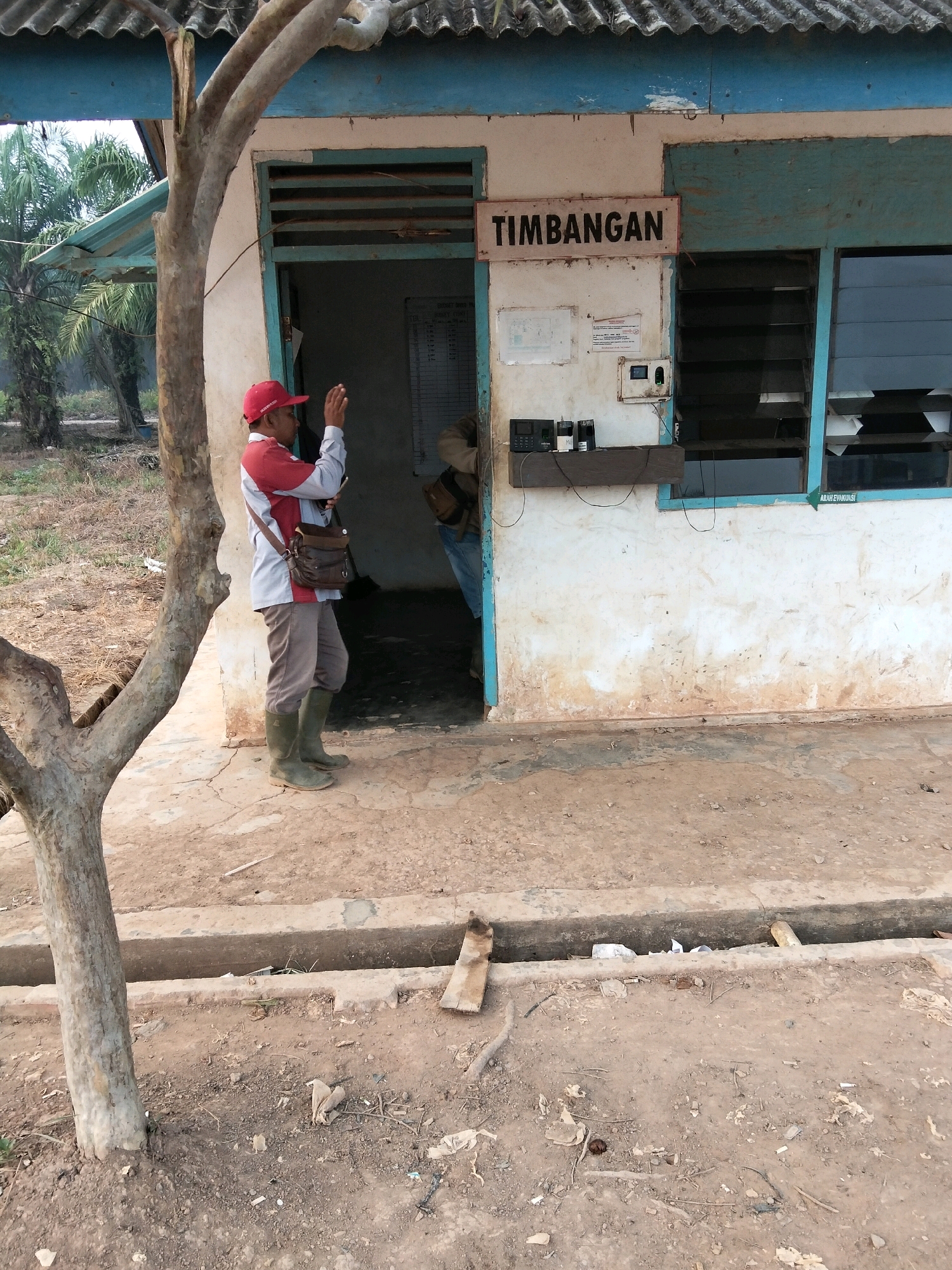 A man wearing a red cap, a red and gray shirt, khaki pants, and green rubber boots stands outside a white building with a blue trim. He is holding a phone up to his face and appears to be taking a picture. A brown bag is slung across his shoulder. The building has a sign above the entrance that reads "Timbangan". Inside the building, partially visible through the doorway, a person in a denim jacket and jeans is seen wearing a backpack. Electrical equipment, including what appears to be a scanner and two other devices, is mounted on the wall near the entrance. The ground outside the building is dirt and concrete. A tree trunk is visible in the foreground to the left of the man. The background shows palm trees and dry vegetation. The scene appears to be in Teluk Tenggirik, Indonesia.