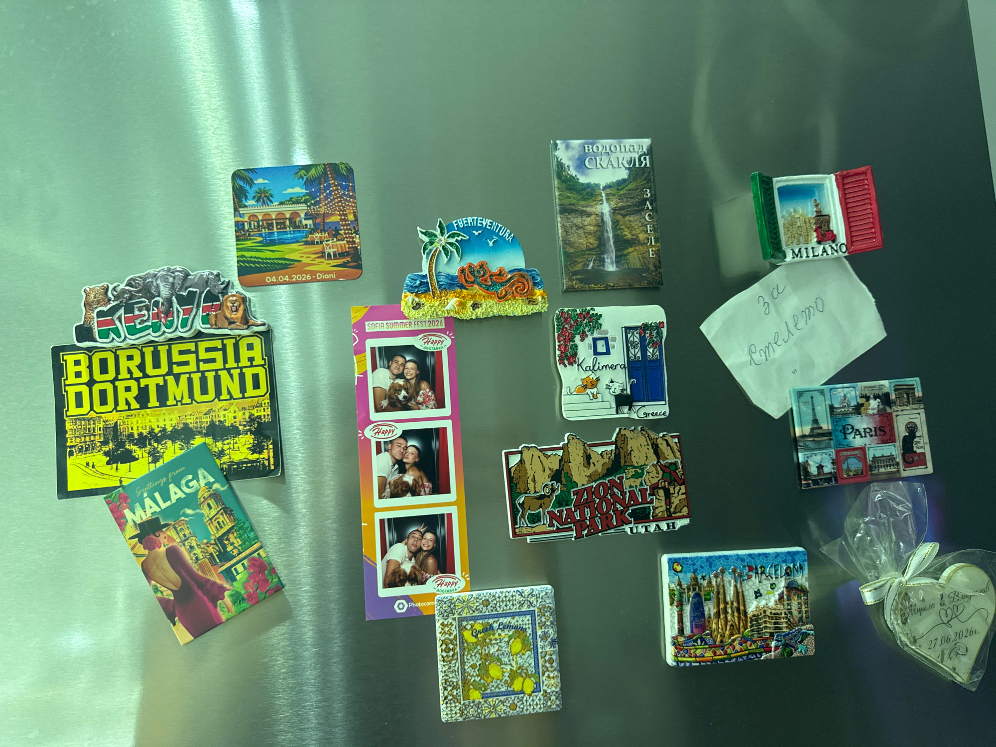 A collection of refrigerator magnets are affixed to a stainless steel refrigerator door. The magnets depict various locations and designs. One magnet shows a resort with palm trees and a pool, dated "04.04.2026 - Diani". Another magnet features a waterfall with text in Cyrillic, which translates to "Skaklia Waterfall". A magnet with the text "FUERTEVENTURA" depicts a palm tree and a beach scene. A magnet for "BORUSSIA DORTMUND" is also visible, along with a magnet for "MALAGA". There is a photo strip of three pairs of people, with "SOFIA SUMMER FEST 2024" written at the top. Another magnet displays a Greek island scene with "KALIMERA" and "GREECE". A magnet for "ZION NATIONAL PARK UTAH" shows a desert landscape with an animal. A magnet for "PARIS" shows various Parisian landmarks. A magnet for "BARCELONA" depicts the Sagrada Familia. Additionally, there is a heart-shaped magnet with handwritten text that appears to be a date and names, and some other magnets with tile-like patterns. The overall environment is an indoor setting, likely a kitchen, given the refrigerator.