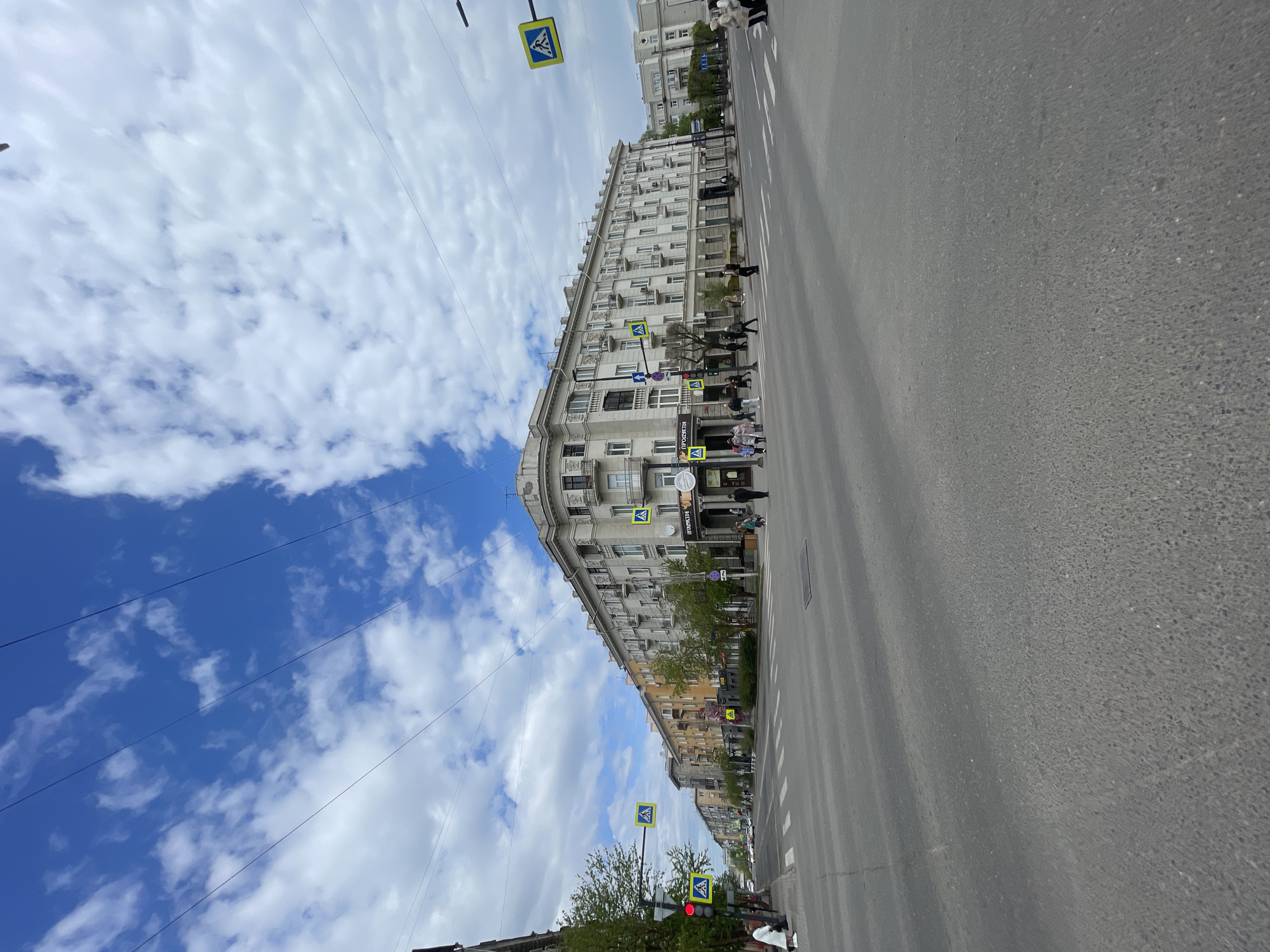 The image captures a street scene in Krasnoyarsk, Russia. The perspective is from a low angle on the road, looking towards a tall building with a curved facade on the right side of the frame. The building appears to be multi-storied, with numerous windows and architectural details.
Several pedestrians are visible along the sidewalk in front of the building, engaged in walking. Pedestrian crossing signs are situated along the street, indicating designated crosswalks.
The road occupies a significant portion of the frame. Overhead power lines stretch across the sky.
The sky is predominantly blue with scattered white clouds.