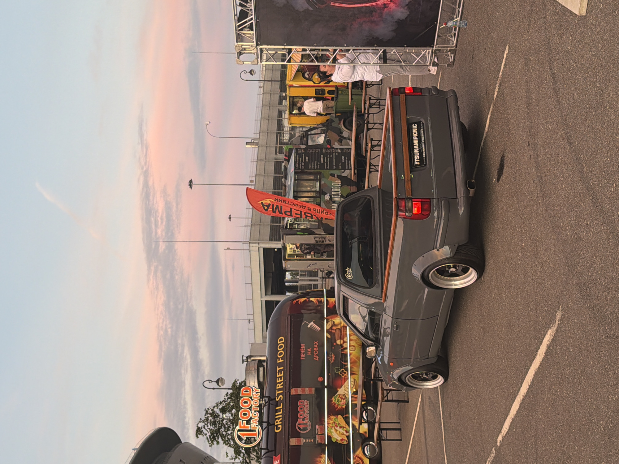 An outdoor scene depicts a food truck event at dusk, with a prominent grey pickup truck in the foreground. The truck, a modified smaller model, faces away from the viewer, showcasing its wide fender flares, custom wheels, and wooden trim on the tailgate. Behind the truck, several food trucks are parked, including a "1 Food Factory" truck with colorful graphics of food items and a camouflage-patterned truck with menu boards. A red banner with Cyrillic lettering is displayed near the camouflage truck. People are visible interacting with the food trucks, with one person standing near a yellow truck and another near a large display structure on the right side of the image. The environment suggests a public gathering space, likely a parking lot or open area, with overhead lighting and a cloudy sky displaying hues of pink and blue from the setting sun.