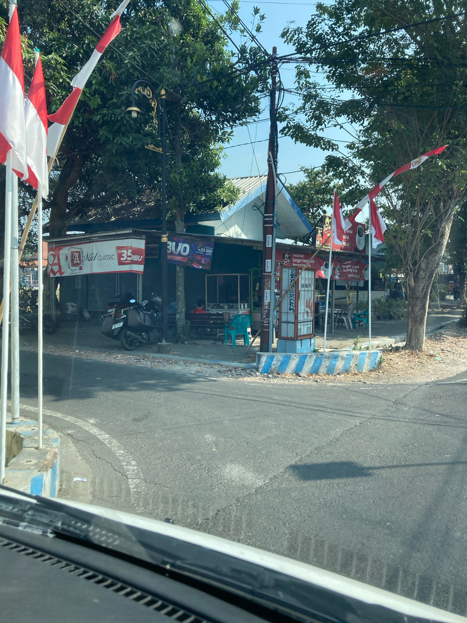 In the city of Unknown, Unknown, an exterior shot captures a street-level view of a building with a covered outdoor seating area, seen from the perspective of someone inside a vehicle. Red and white Indonesian flags are prominently displayed on flagpoles in front of the building. The building appears to be a small shop or eatery, with advertisements visible on its facade, including one for "Mild Classic 35 iBU" and another featuring the word "BUO" with a price. A motorcycle is parked in front of the building, and several chairs are visible in the seating area. Trees line the street, and power lines are strung overhead. The scene is bathed in bright sunlight, casting shadows on the asphalt road.