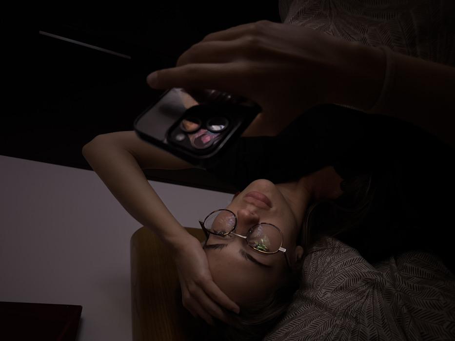 A woman in glasses is lying down, holding her head with one hand. She has a phone in her other hand, held above her. The scene is dimly lit. The woman is wearing a dark shirt and lying on a patterned surface. The camera lenses of the phone are visible and reflect the light.