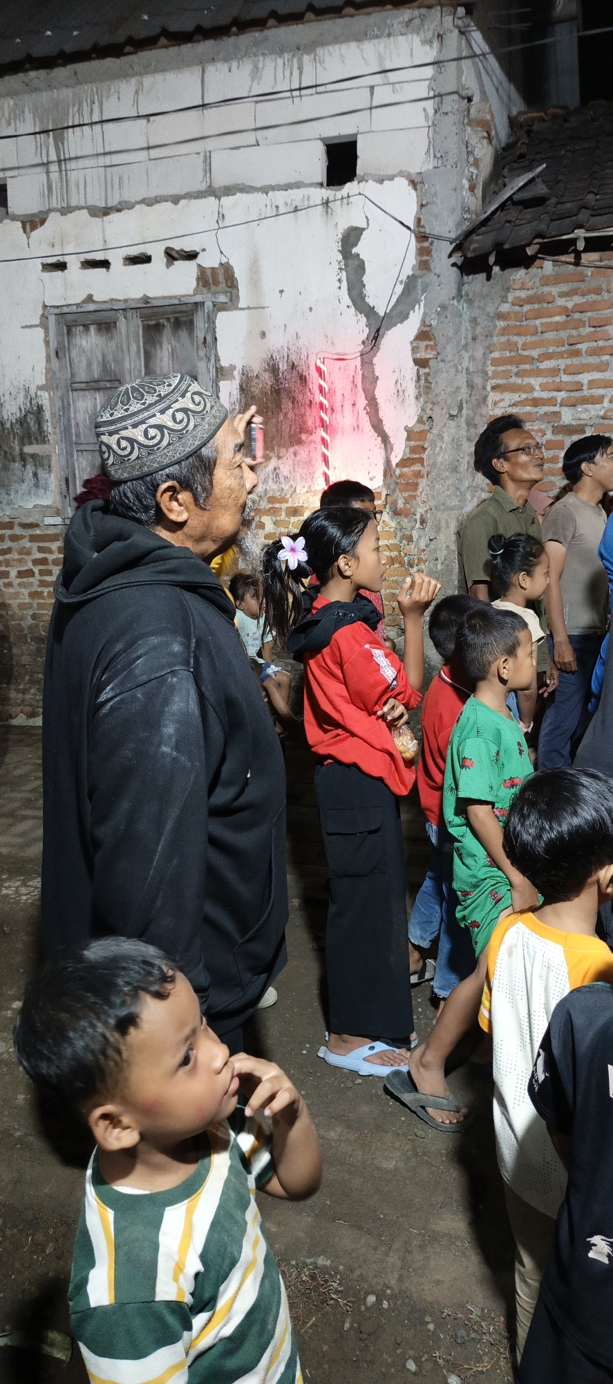 A group of people, including adults and children, are gathered outdoors at night. The background appears to be a brick building with a decaying facade and a wooden window. A man in the foreground, wearing a black hoodie and a patterned cap, is holding up a mobile phone, possibly taking a picture or recording. To his right, a young girl in a red top and black pants looks upwards, a pink flower in her hair. Other individuals are visible further back, some looking in the same direction as the girl. The lighting suggests artificial light sources are present, casting shadows and illuminating the scene. The ground is uneven and appears to be dirt or gravel.