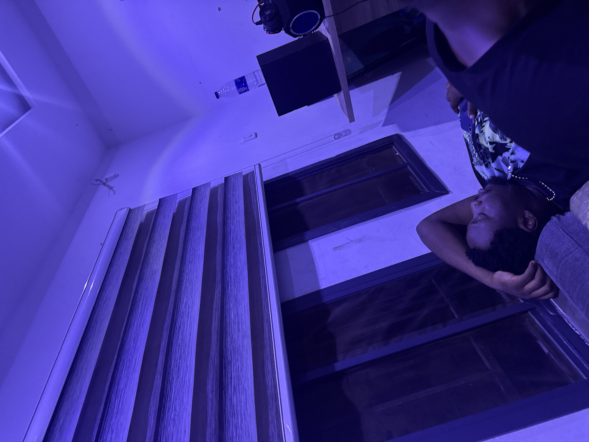 A low-angle, blue-toned photograph depicts a domestic interior, possibly in Abuja, Nigeria, although no specific landmarks confirm this. In the foreground, a dark-skinned person lies on a couch. Above and to the left of the individual are two closed windows. Blinds obscure a portion of one window. Above the window is a shelf with a black speaker. A water bottle is next to the speaker. Ceiling details and a small, stylized airplane decoration complete the scene.