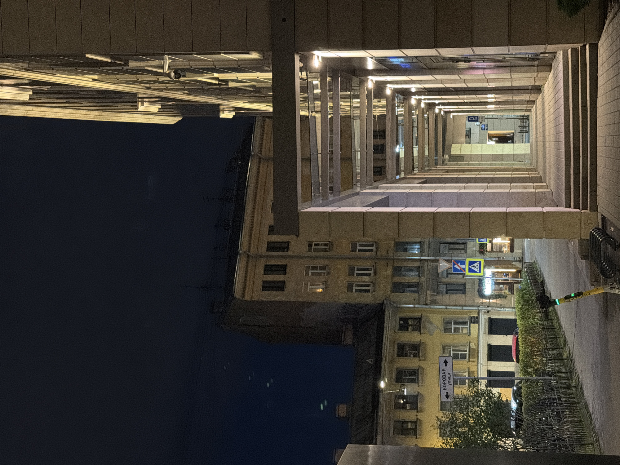 The image depicts an outdoor urban scene at night in Saint Petersburg, Russia. A covered walkway with a modern, illuminated ceiling extends into the distance on the right side of the frame, flanked by a light-colored stone building. The walkway features steps leading up from the foreground.
To the left of the walkway, a paved path runs alongside a metal fence. Beyond the fence, there is a small tree, a bush, and a yellow scooter with a green light. Further in the background, there are several older, multi-story buildings with visible windows, some illuminated. A street sign with Russian text and arrows pointing left and right is mounted on a pole in front of these buildings. Traffic signs, including a pedestrian crossing sign and a blue circular sign, are also visible.
Several vehicles are parked along the street, and a red object, possibly the roof of a car, is visible behind the bush. The sky is dark blue, indicating nighttime. The overall impression is a contrast between modern architecture and older buildings in an urban setting.