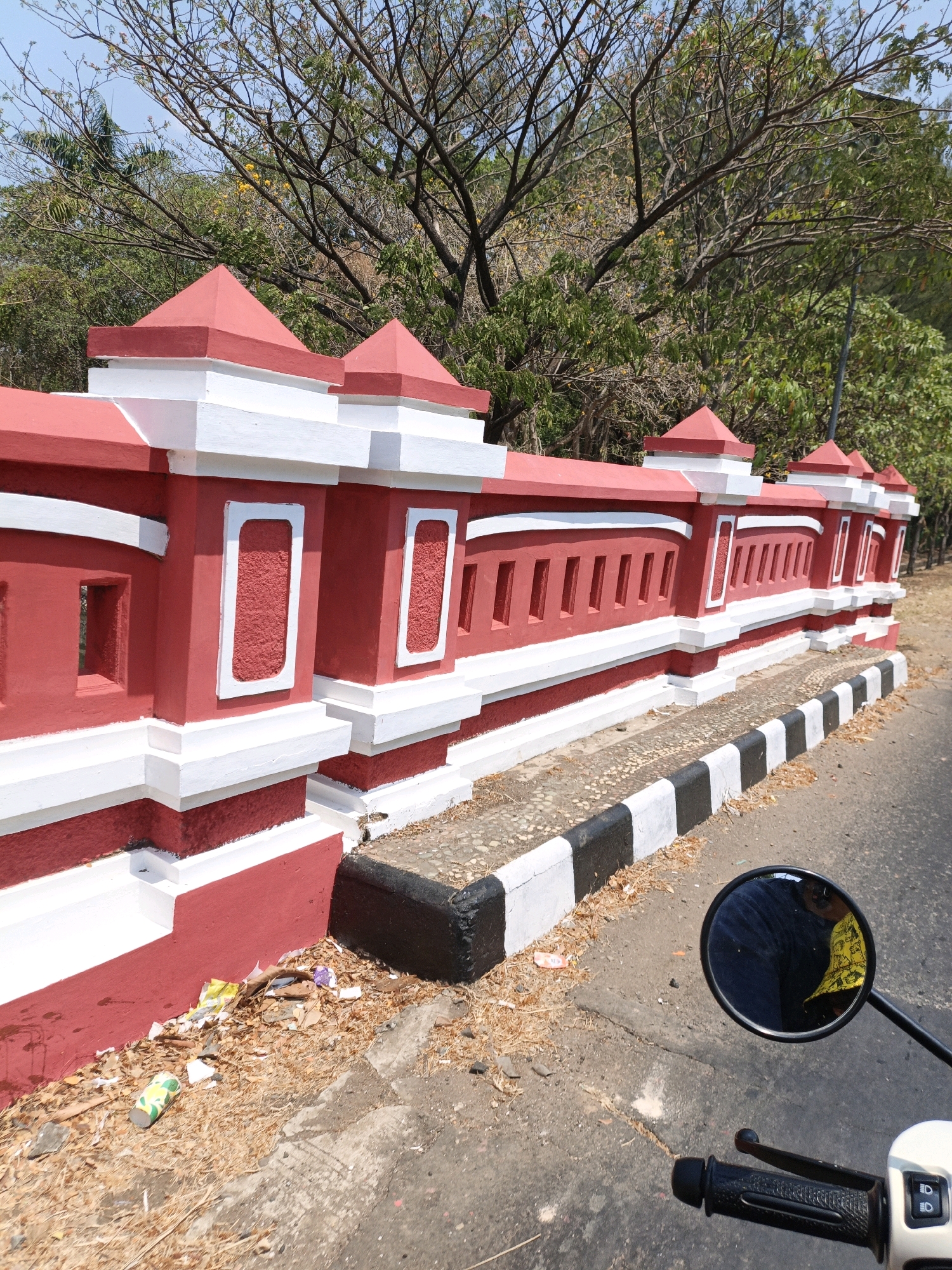 This is an eye-level, medium shot of a red and white concrete barrier along a road in Jepara, Indonesia. The barrier features a decorative design with white accents and pointed finials. The ground beside the barrier is covered in dry leaves and some scattered litter, including plastic cups and wrappers. A black and white striped curb runs along the edge of the road. In the foreground, a motorcycle handlebar and a round rearview mirror are visible. The mirror reflects the image of a person wearing a yellow shirt. Trees with sparse branches and some green foliage are in the background. The sky is clear and blue.