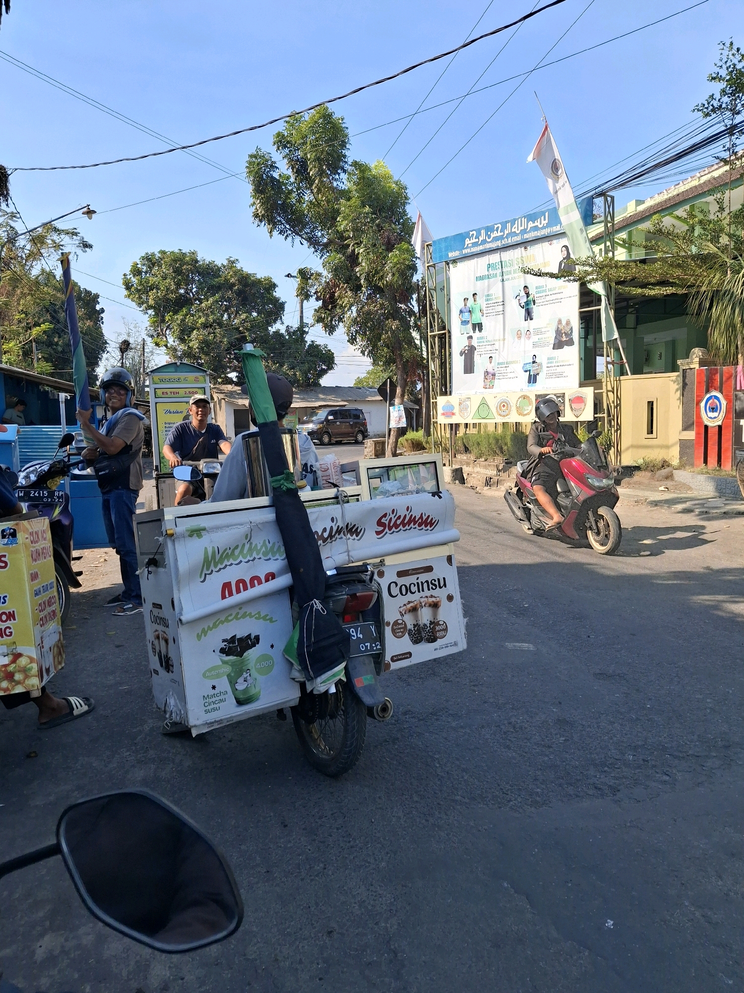 In Lumajang, Indonesia, on a sunny day, a street scene unfolds with several food and drink vendors. In the foreground, a motorcycle with a sidecar serves as a mobile beverage stand, advertising "Cocinsu" and "Macinsu" with prices listed. Another vendor with a similar setup is partially visible on the left, displaying "CLOK CLOK" and "IKAN" on its signage. Two men are associated with these vendors; one, wearing a helmet, stands by the motorcycle on the left holding a green umbrella-like object, while another man in a dark shirt and helmet rides a motorcycle that is parked or moving slowly. Further down the street, a woman on a red scooter drives past. In the background, a large banner with Arabic calligraphy and Indonesian text, likely advertising a school or institution, is displayed. The environment is typical of a bustling street with trees, power lines, and buildings lining the road. The time appears to be around 07:22 based on the visible license plate.