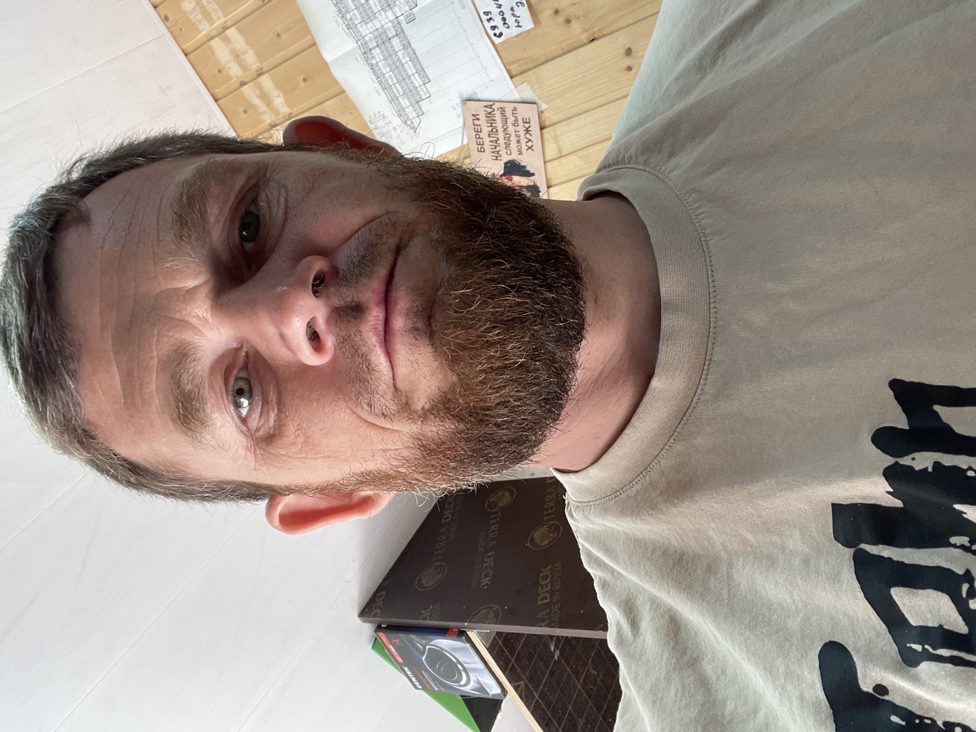 A man with brown hair and a beard is looking directly into the camera, with a slight frown on his face. He is wearing a light-colored t-shirt with a dark, bold graphic design. The background appears to be a room with wooden paneling on the right wall, and what looks like shelves or storage units on the left. There are also some papers or documents visible on the wall behind him. The overall lighting is natural, suggesting it's daytime. There is no specific indication of this being in Krasnodar, Russia, based solely on the visual information provided.