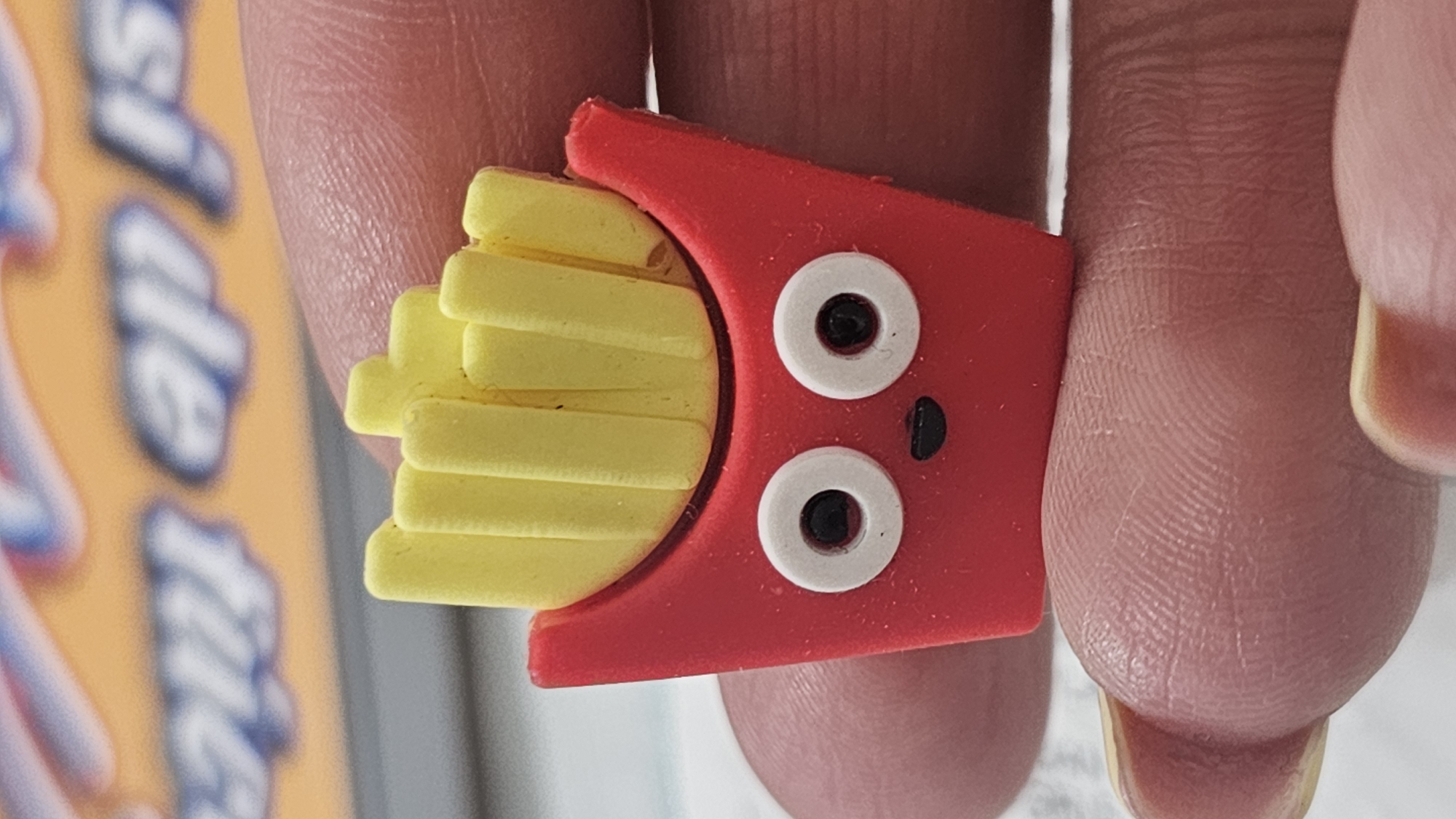 A close-up photograph shows a red, cartoon-style French fry eraser with a face, held between two fingers. The eraser has yellow fries protruding from the top of the red container, and a face with two white eyes and a black dot for a mouth. The background is out of focus but appears to include a box with text in blue and orange. There is no indication in the image that it was taken in San Bernardino Tlaxcalancingo, Mexico.