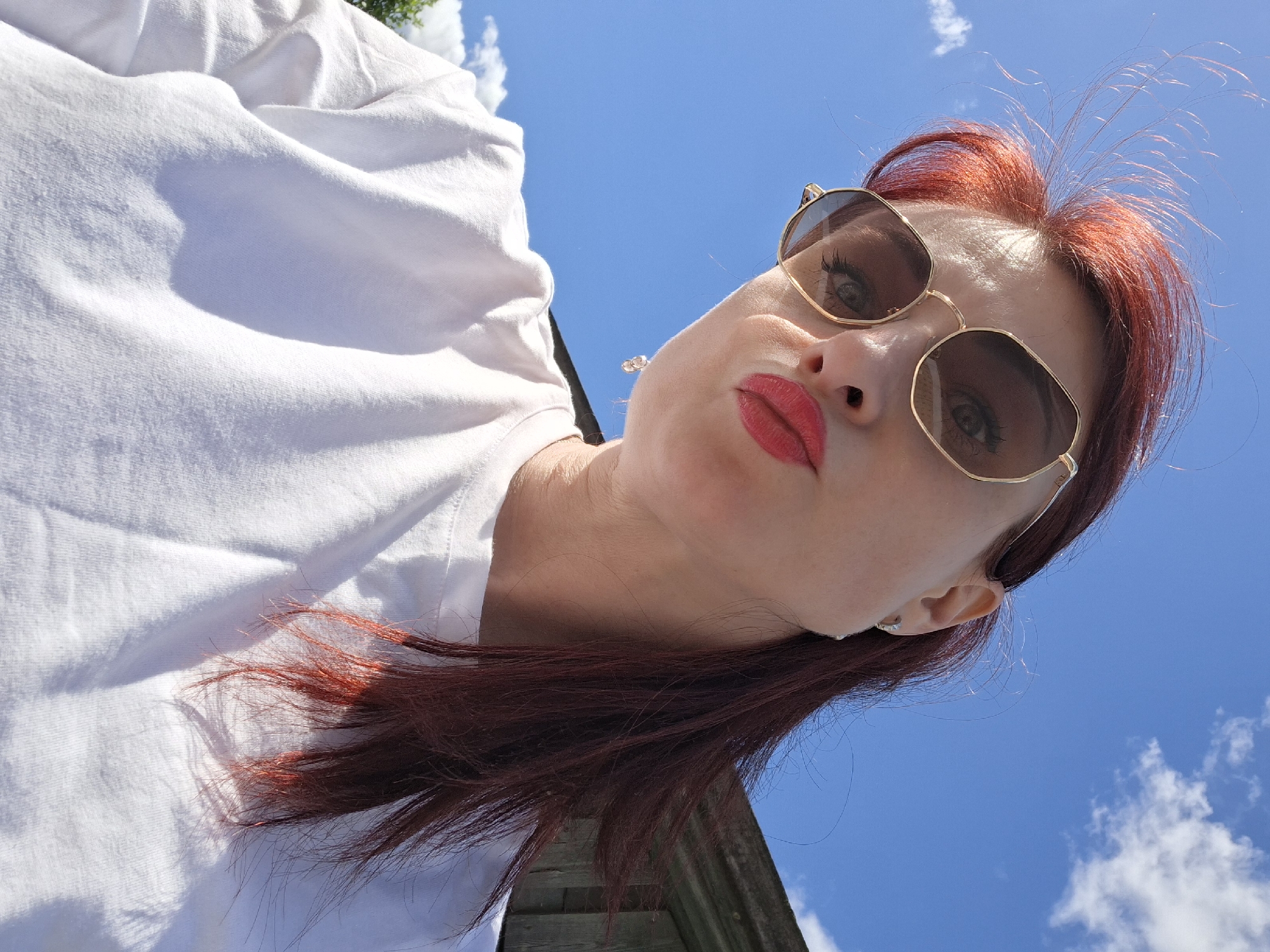 A low-angle, upward-facing photograph shows a woman with red hair, wearing sunglasses and bright red lipstick, pouting. She is wearing a white t-shirt, with bright blue sky and scattered white clouds visible behind her. There's a hint of wooden structure visible at the very bottom edge of the frame. The picture seems to be taken outdoors under bright daylight conditions. The city of Смалявічы, Belarus is not discernible from the given visual information.