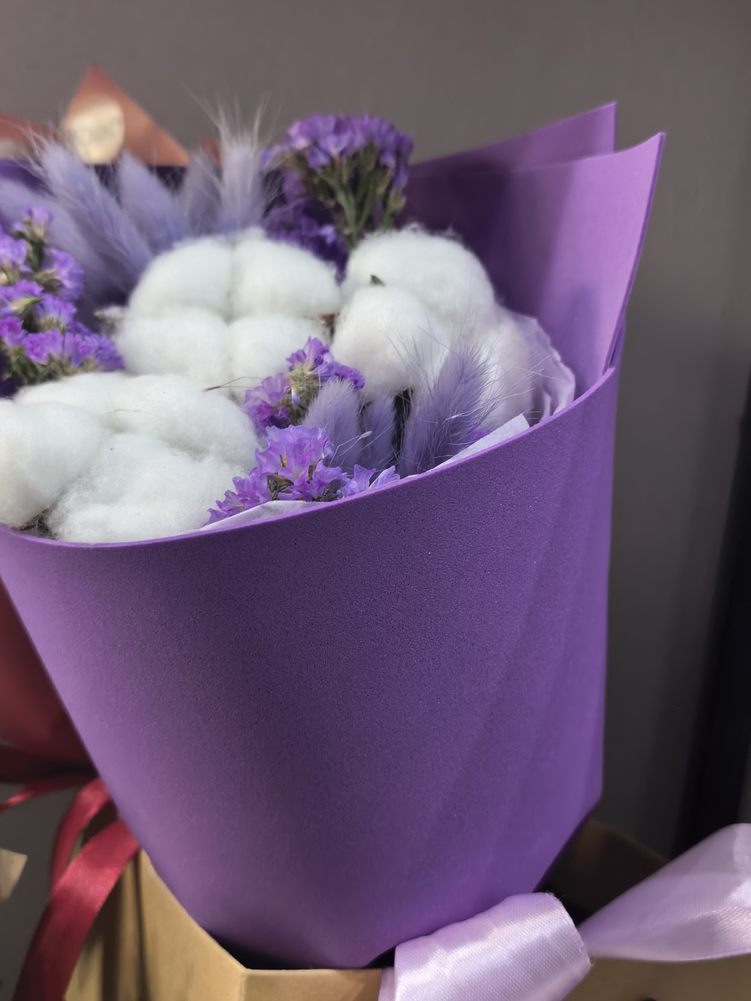 A close-up photograph depicts a floral arrangement, specifically a bouquet, set in an indoor environment. The bouquet is predominantly wrapped in a matte, dark purple material, possibly foam or textured paper, which exhibits a fine, granular surface. The upper edges of the wrapper are curved and folded inwards, revealing the bouquet's contents.
Within the purple wrapper, multiple fluffy, spherical white objects resembling cotton balls are prominently featured. Interspersed among these white elements are clusters of small, dried or preserved purple flowers, consistent with Limonium (Statice), providing textural contrast and color. Additionally, several slender, light purple, feathery plumes are visible, adding a delicate, soft dimension to the arrangement.
The bouquet rests within a partially visible, light brown or cardboard-colored container or bag, seen at the lower edge of the frame. Two distinct ribbons are associated with the presentation: a deep red or burgundy satin-like ribbon is partially visible on the left side, while a lighter, pale lavender satin-like ribbon, appearing to form a loose loop or knot, is present on the right.
The background is indistinct and out of focus, consisting of muted, solid colors, primarily dark grey on the right and a slightly lighter, brownish-grey on the left, indicating an interior setting. The lighting is soft and even, illuminating the textures and colors of the bouquet without casting harsh shadows. The specific location in the city of Unknown, Unknown, cannot be determined from the image.
