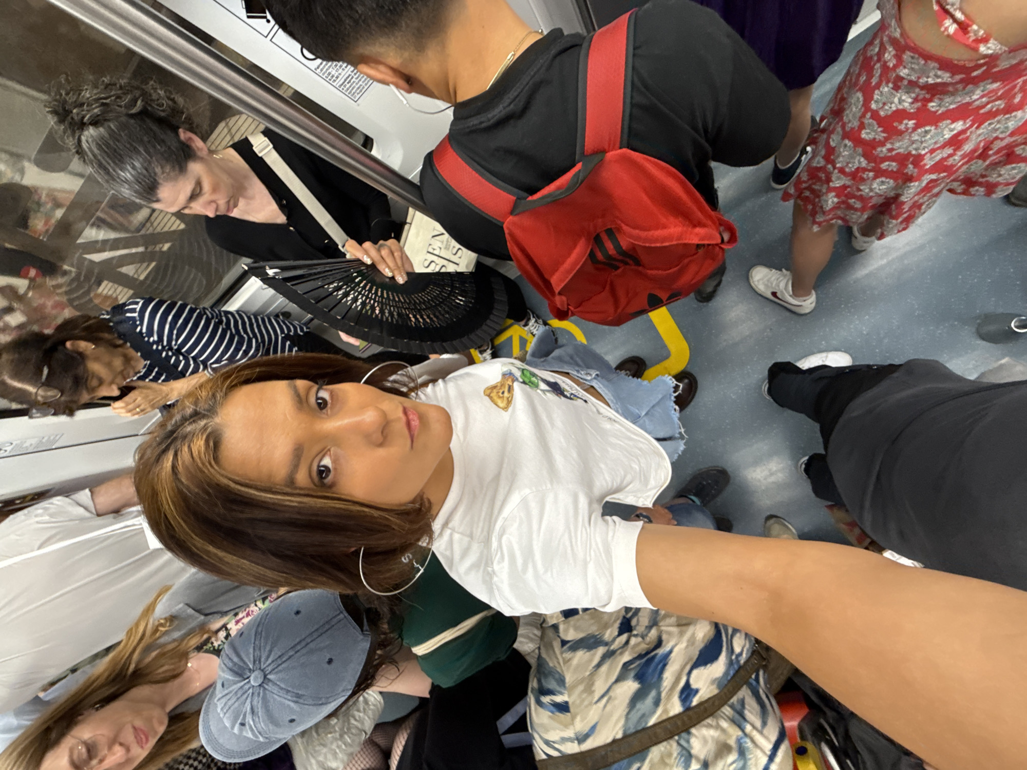 The image depicts a crowded interior of a Barcelona Metro train carriage. Several passengers are visible, with the foreground dominated by a person taking a selfie, showing their arm and face. To the left, a woman wearing glasses and a grey jacket is looking down. Behind her, another woman in a blue and white striped shirt is also looking down. In the middle-right, a woman with her hair in a high bun holds a black fan. To her right, a man wearing a black hoodie with red stripes on the sleeves and black shorts has a red bag slung over his shoulder. Further back, other passengers' legs and feet are visible, indicating a densely populated environment. The floor of the train carriage is a light blue with yellow markings. The overall scene is indicative of rush hour or a busy travel period on public transport within Barcelona.