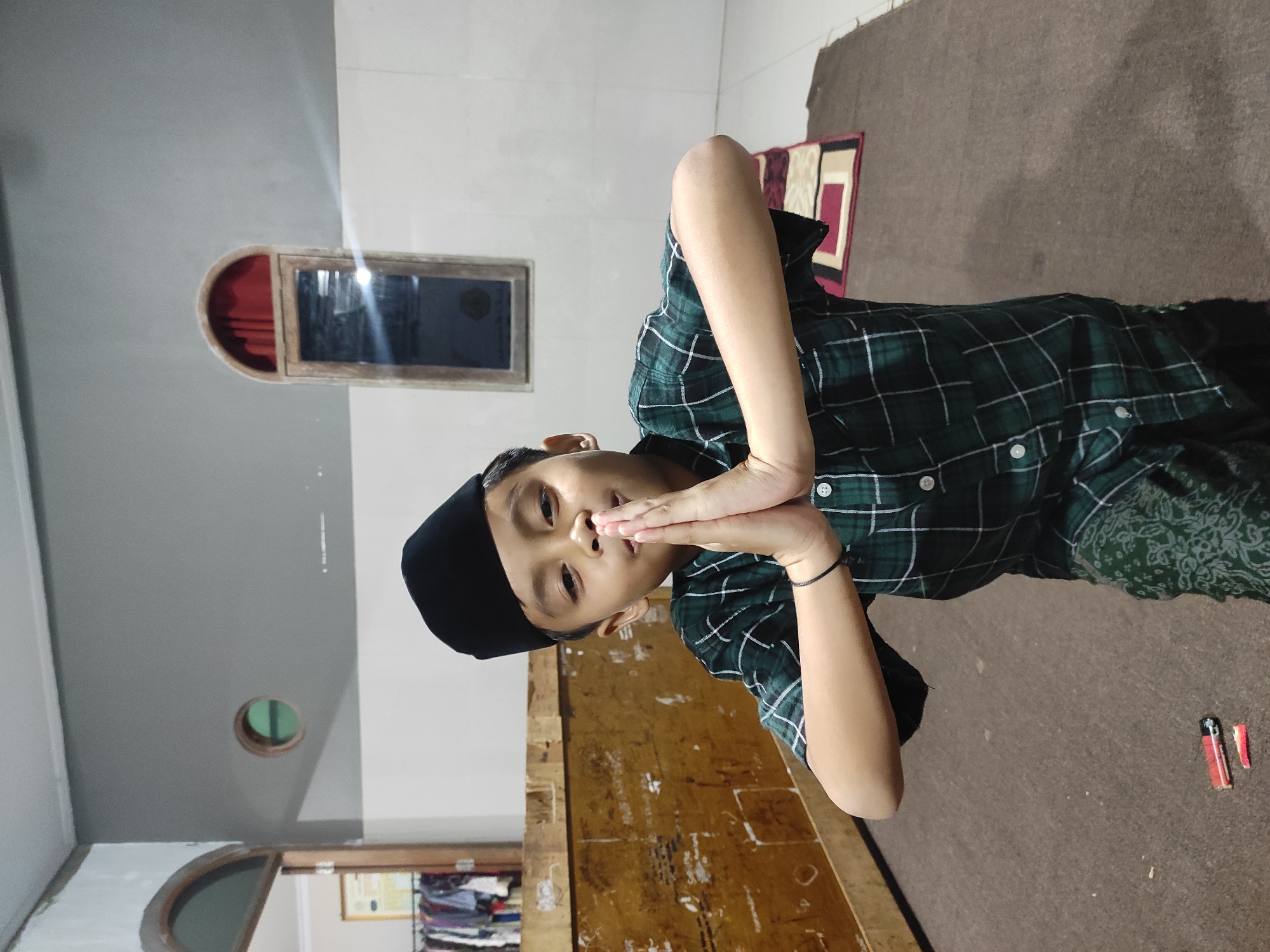 A young boy wearing a black peci and a green plaid shirt stands on a brown carpet. His hands are clasped together in front of his chest, palms touching. He is in a room with light-colored walls and a grey ceiling. In the background, there is a wooden structure, a small circular window, and a tall, narrow window with red curtains. The boy is positioned in the foreground of the image, and the background is somewhat out of focus. The lighting appears to be artificial and casts shadows on the floor. There is a small red and black object on the carpet in the lower left corner of the image. The scene is indoors, and based on the cultural attire and environment, it is likely a residential setting in Indonesia.