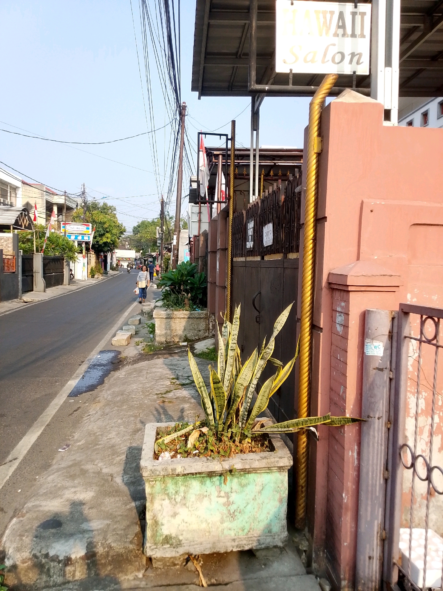 The image captures a street scene in East Jakarta, Indonesia, bathed in the late afternoon sun. The road, marked with a white line, is bordered by a concrete sidewalk. A large, rectangular planter with a snake plant is positioned on the sidewalk, showing signs of wear and discoloration. Across the street, buildings with red roofs and white walls are visible, along with trees and utility poles supporting a tangle of overhead wires. A sign reading "HAWAII Salon" is mounted above a rusted metal gate. Further down the street, a woman walks away from the camera, carrying a bag. Several other individuals and a motorcycle are visible in the distance. The overall impression is of a typical urban neighborhood in Indonesia.