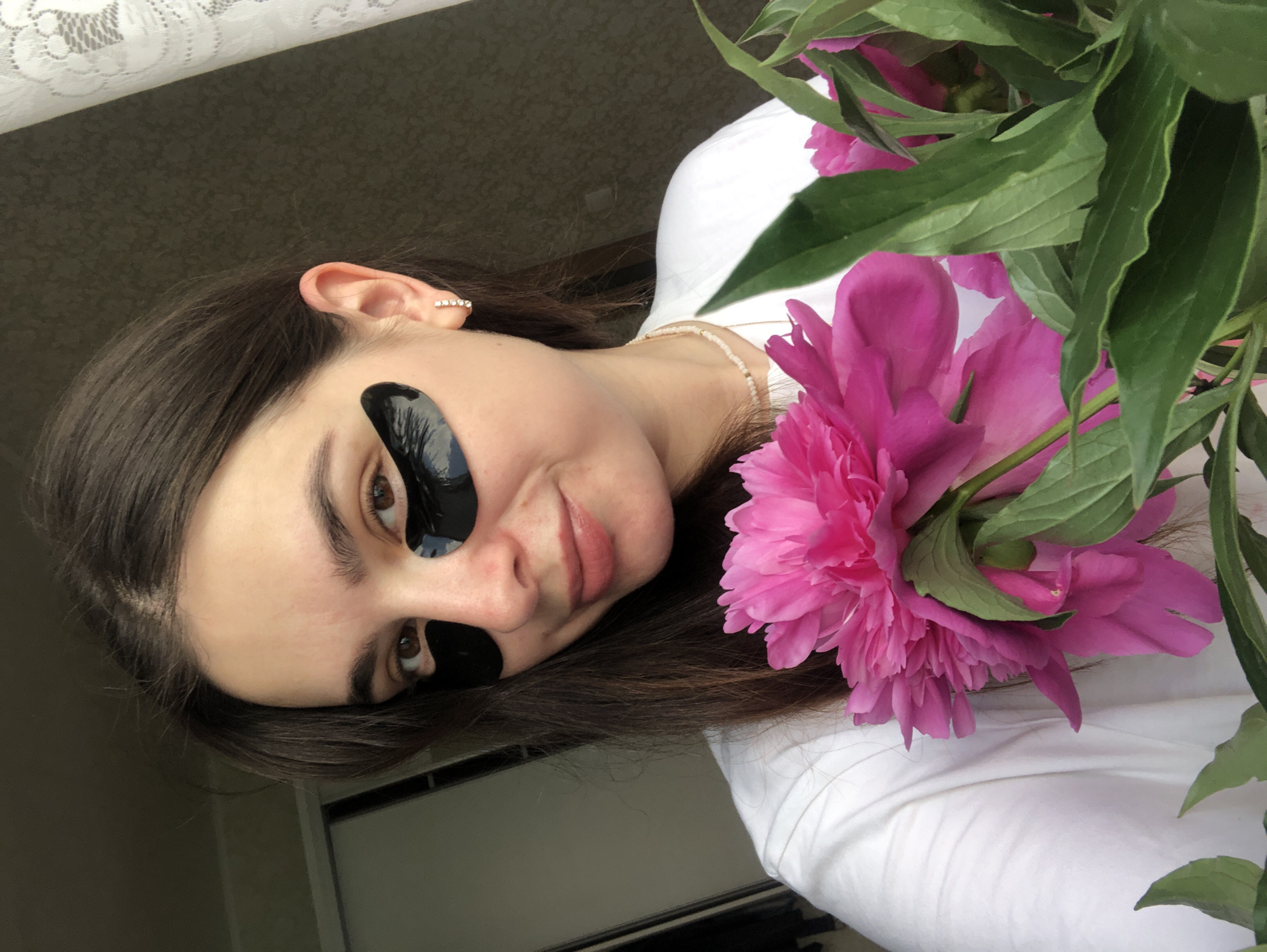A young woman with dark brown hair is taking a selfie indoors, holding a bouquet of bright pink peonies in front of her white t-shirt. She is wearing black under-eye patches, a simple necklace, and small hoop earrings. The background is indistinct, featuring a dark wall and a section of white lace curtain. The lighting suggests it is daytime. The location is not specified in the image and cannot be determined.