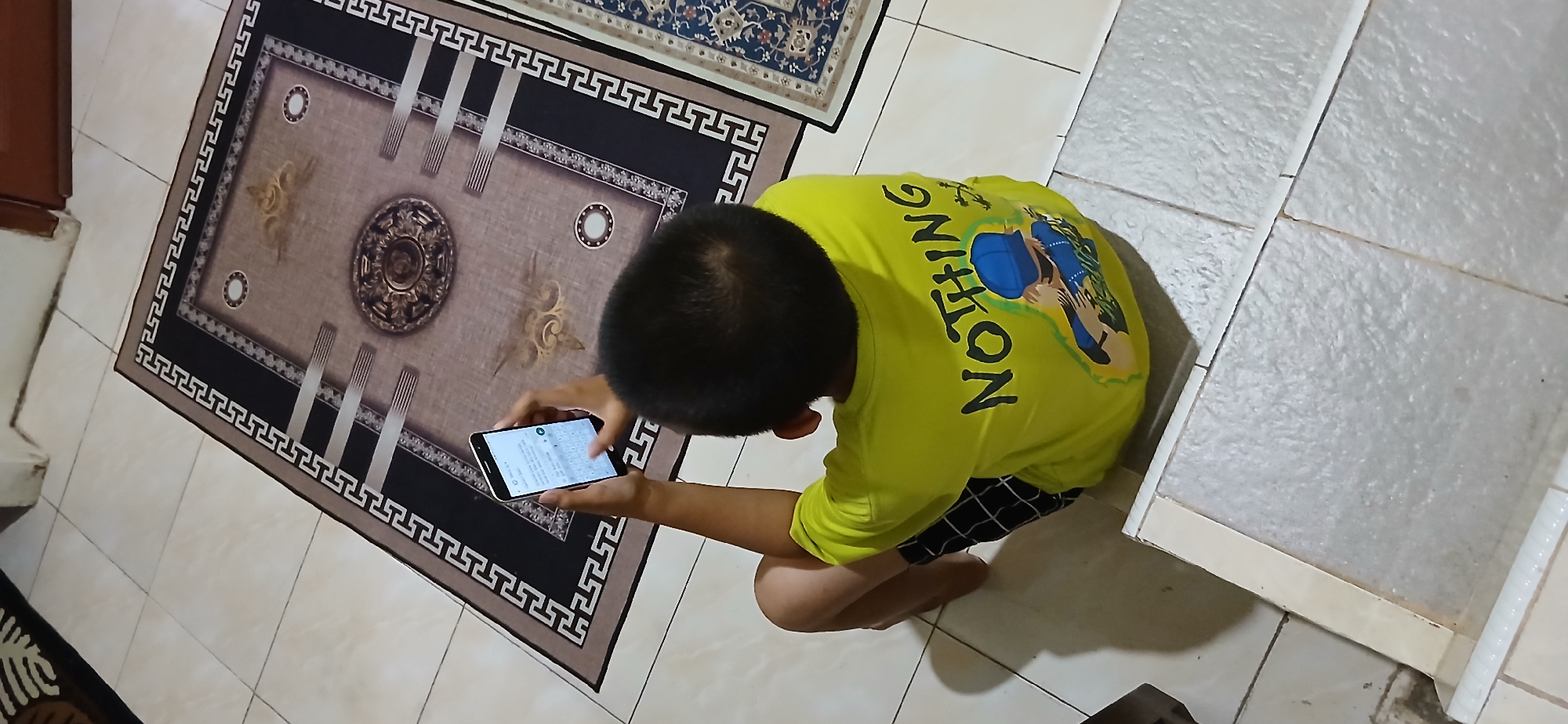 A child is sitting on a tiled floor, facing away from the camera. The child is wearing a bright yellow t-shirt with the word "NOTHING" printed in black lettering across the back. Below the word, there is an illustration of a person wearing a blue cap and jacket. The child is holding a smartphone in their hands, looking down at the screen. The background shows two rugs on the floor. One is a large, ornate rug with a dark border and a central medallion design. To the right, a portion of another rug with a blue pattern is visible. The setting appears to be an indoor residential space in Samarinda, Indonesia, likely a living room or a common area, given the tiled flooring and rugs. The lighting suggests it is daytime, possibly with artificial light supplementing natural light.