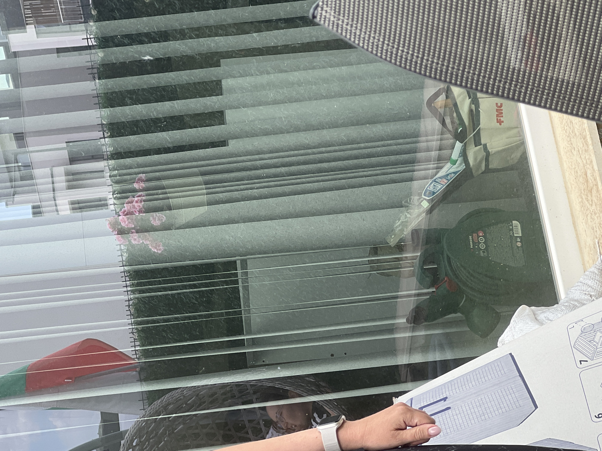 The image, taken from an exterior perspective looking into a glass-covered area, shows a reflective scene composed of a green and red flag, pink flowers in a pot, a dark wicker chair, and a portion of a person's arm and hand wearing a white smartwatch. The background is dominated by vertical, pale green blinds and a building with multiple windows.
Inside the glass-covered area, several objects are visible. A dark green, cylindrical object with red accents, identified by the "PARKSIDE" logo, appears to be a pump or similar gardening equipment. Next to it is a light-colored canvas bag with red "FMC" lettering, containing a white and green object wrapped in plastic, likely a tool or accessory. The overall environment suggests an outdoor or semi-outdoor space, possibly a patio or garden area adjacent to a building. The scene is illuminated by natural light, creating reflections on the glass that obscure some details and blend the interior and exterior elements.