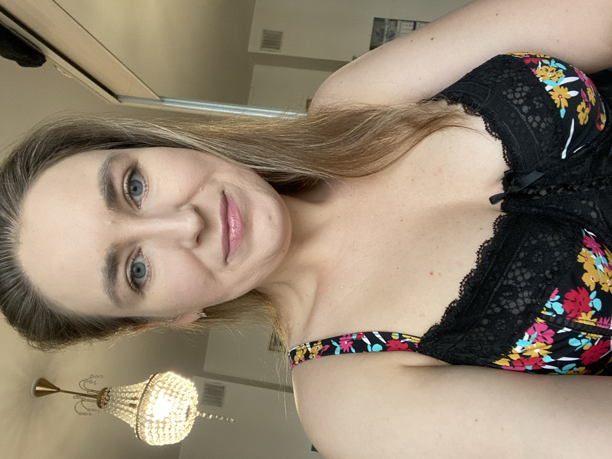A woman with fair skin and light brown hair is taking a selfie. She has blue eyes, and her lips are glossy. She is wearing a black bra with a floral pattern and lace trim. Behind her is a wall and a chandelier. The location cannot be determined from this image.