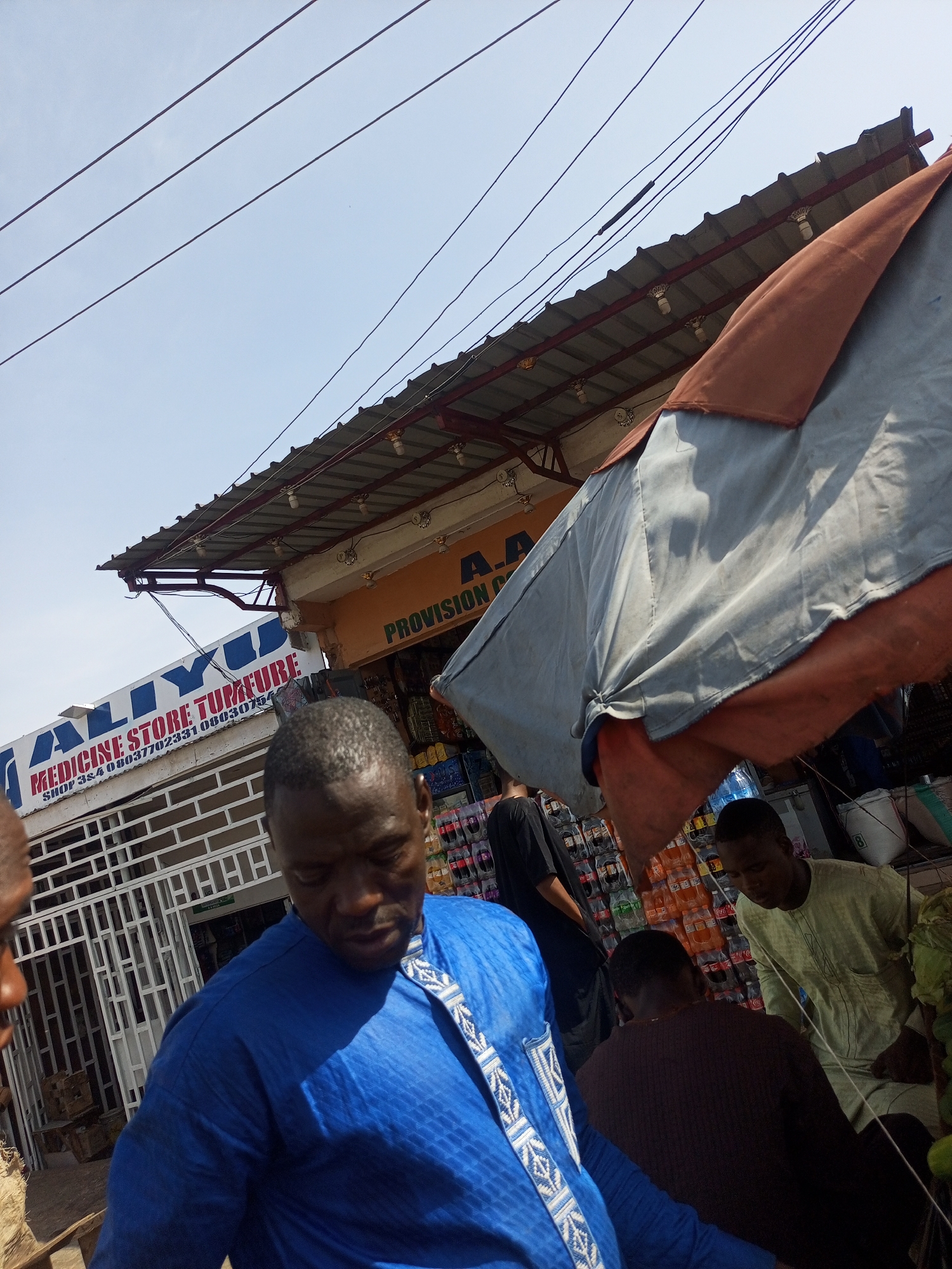 The image captures a vibrant outdoor market or street scene in Tempure, Nigeria, under a clear, bright blue sky. Harsh sunlight and distinct shadows indicate it is likely midday or early afternoon.
In the foreground, a man wearing a bright blue traditional attire with white geometric patterns looks downwards. His face and head are prominent, with short dark hair. To his left, another person's head is partially visible.
Behind the foreground figures, the scene reveals several commercial establishments. A shop with a white security grate features a large blue sign that reads "ALIYU MEDICINE STORE TUMFURE." Below this, contact details are visible: "SHOP 324 08037702331 0803075...". Adjacent to it, a yellow-orange storefront displays the partial text "PROVISION A.A."
A large, multi-colored awning or umbrella, primarily grey with rust-orange accents, stretches across the midground, providing shade for market activities. Beneath this canopy, several individuals are present. One person in a black top stands with their back to the viewer, while another in a light green traditional garment looks down, possibly arranging goods. A third person, seen from behind, wears a dark brown top. Stacks of bottled beverages, including soft drinks and water, are visible on shelves in the background, indicating a provision store or stall. Overhead, power lines crisscross the sky. The overall impression is one of an active, bustling market area.
