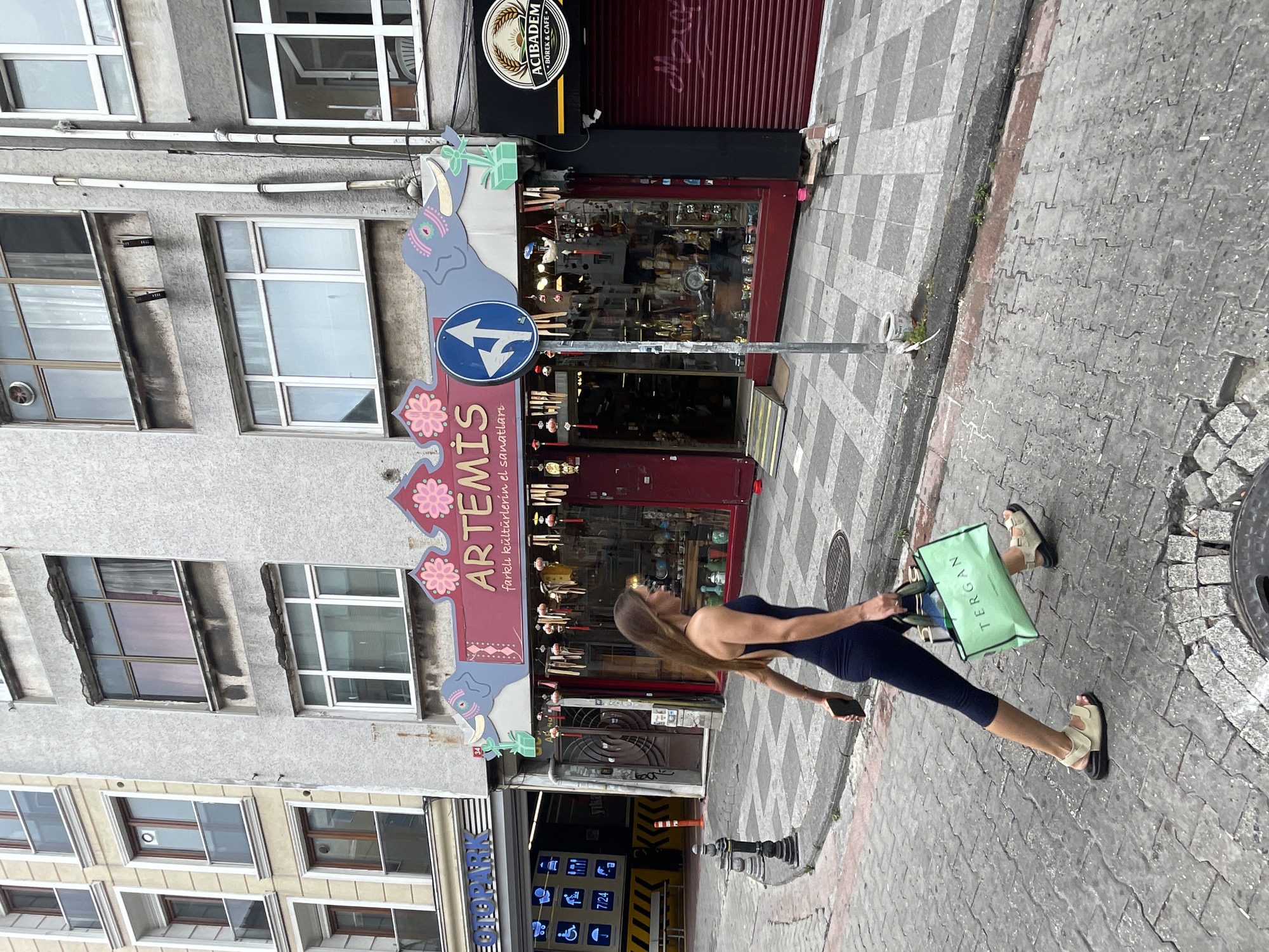 A woman in a dark blue sleeveless dress and tan platform sandals walks on a cobblestone sidewalk in Kadıköy, Turkey. She carries a light green shopping bag with the "TERGAN" logo in black lettering in her left hand and a black smartphone in her right hand. The woman is walking past a shop with a red storefront and large display windows showcasing various items. Above the shop, a sign reads "ARTEMIS" in stylized pink lettering, with decorative elephant motifs on either side. A blue directional sign with two arrows is mounted on a pole in front of the shop. To the right of the Artemis shop, there is a shop with a black awning and a red rolling door. The background shows the facades of multi-story buildings. To the left of the Artemis shop, there is a blue and yellow sign for "OTOPARK" indicating a parking garage. The street is paved with grey interlocking stones and some darker grey square tiles.