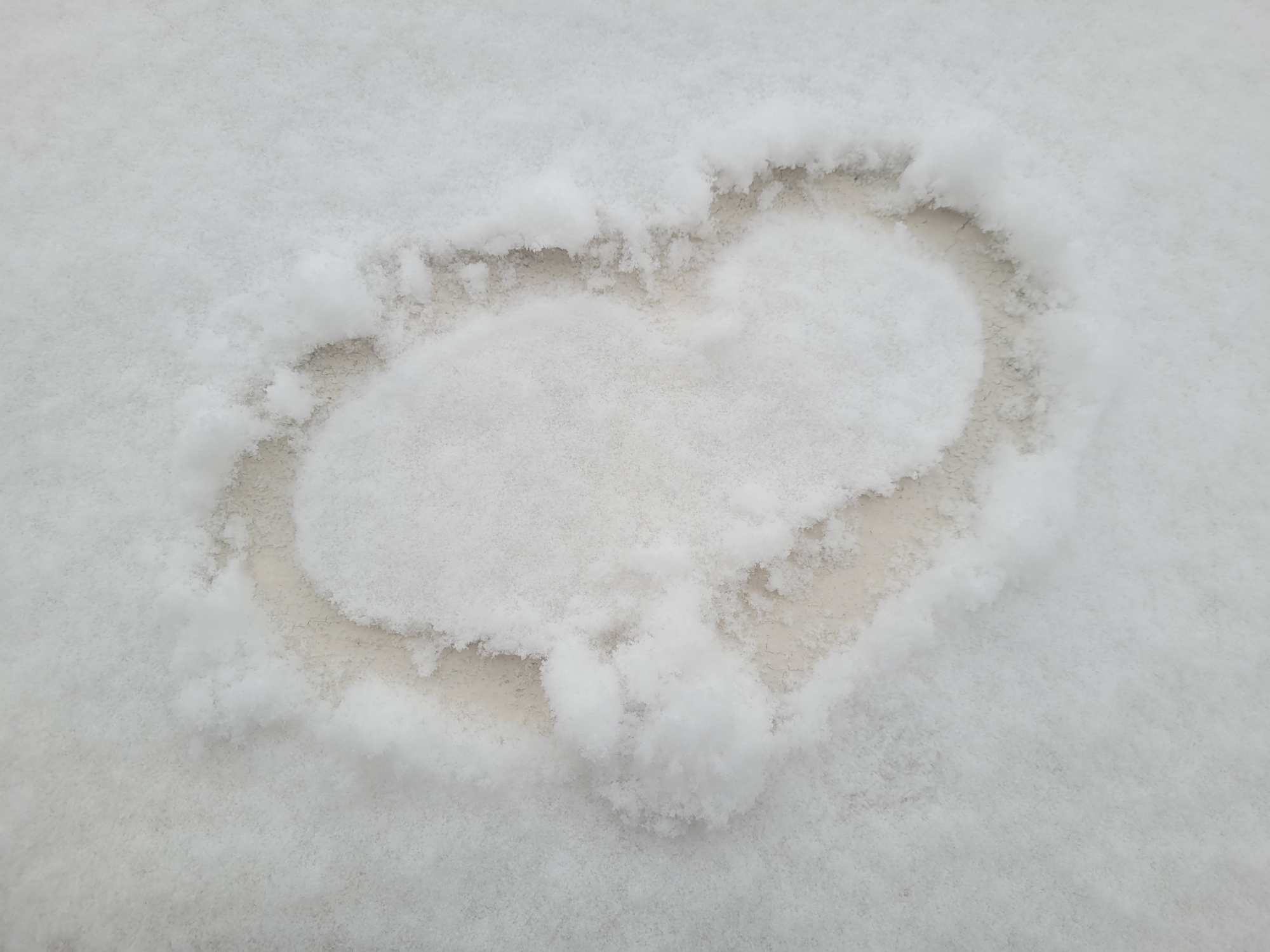 A heart shape has been formed in the snow. The location is not specified in the image and therefore cannot be attributed to Izhevsk, Russia. The background is fully covered in snow except for the inner part of the heart shape where the ground is slightly visible. There are no people, actions, or interactions visible in the image.