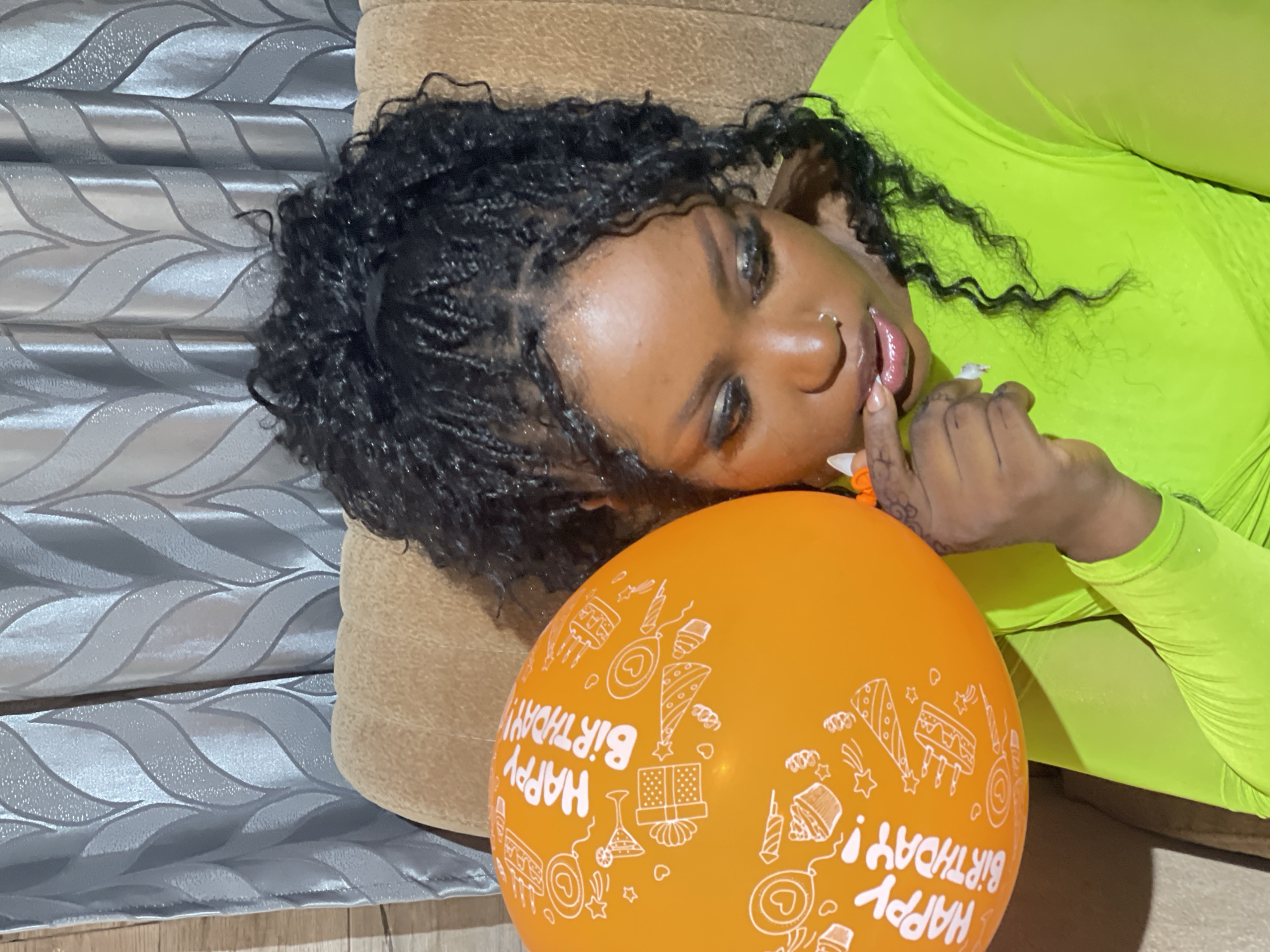 The image features a person, likely female, with dark skin and dark braided hair, lying down on a light brown, upholstered surface, possibly a couch. She wears a vibrant neon green garment and has makeup, including dark eyeshadow and lip gloss. Her expression is calm and direct as she looks towards the camera.
An orange balloon, adorned with white "HAPPY BIRTHDAY!" text and various illustrations of birthday cakes, gifts, party hats, and confetti, is positioned near her head and body. One of her hands, with long, light-colored nails and a ring, is near her mouth. A nose piercing is also visible.
The background shows a decorative wall covering or curtain with a wavy, metallic-like pattern in shades of grey and silver. The scene is set indoors, under artificial lighting, strongly suggesting a birthday celebration or photoshoot. The location context provided is Kuchingoro, Nigeria.