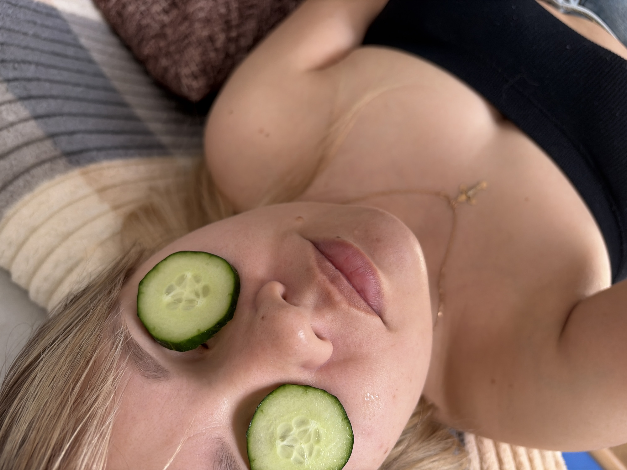 A woman lies on her back with cucumber slices covering her eyes. She has light skin and blonde hair. She is wearing a black top and a gold necklace. She is lying on a patterned blanket with a brown pillow behind her head. The location is undetermined, and there is no indication that it is in Chaikovsky, Russia.
