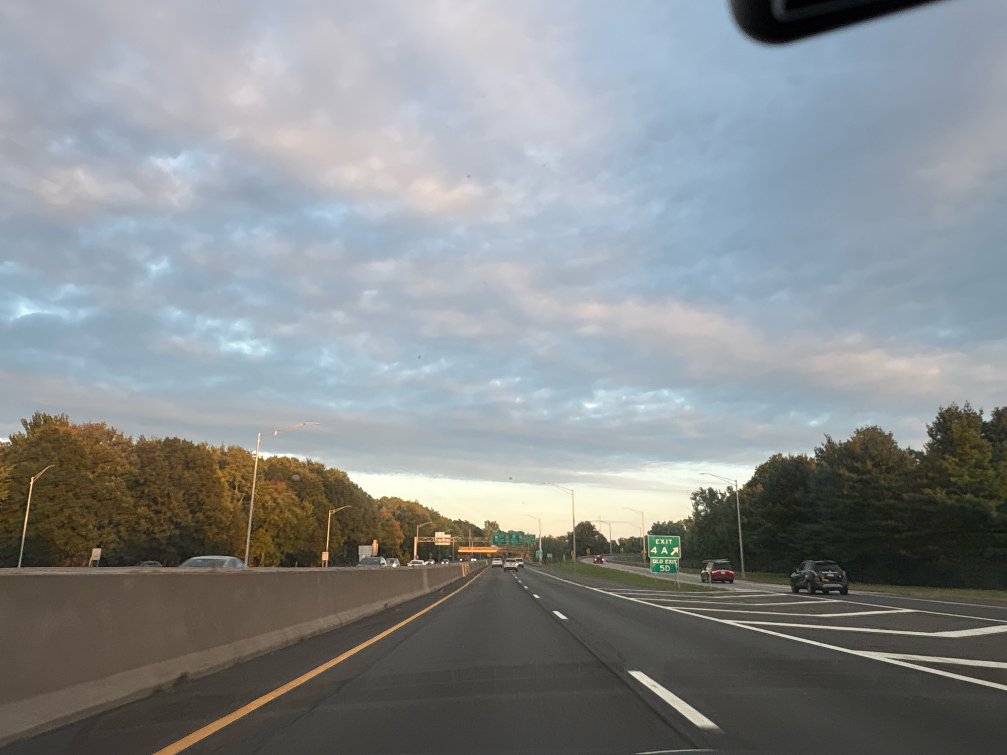 The image shows a highway scene in Glastonbury, United States of America. The highway has multiple lanes and is bordered by trees on both sides. There are several cars on the road, traveling in the same direction. A green exit sign with "EXIT 4 A" and "OLD EXIT 5D" is visible on the right side of the road. The sky is partly cloudy, with the sun setting or rising, casting a warm light on the scene. The overall atmosphere is calm and serene, typical of a suburban or rural highway.