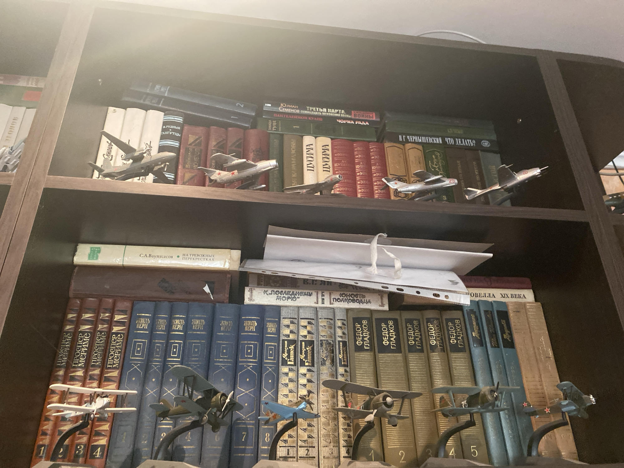 The image shows a close-up view of a bookshelf filled with books and several model airplanes. The bookshelf is dark brown and appears to be made of wood. The books are of various sizes and colors, with some having titles in Russian. The model airplanes are scattered throughout the shelves, some are biplanes and others are jet fighters. The lighting in the image suggests it is indoors, possibly a home or a library. There are no people visible in the image. The environment is cluttered but organized, indicating a collection of books and model airplanes.