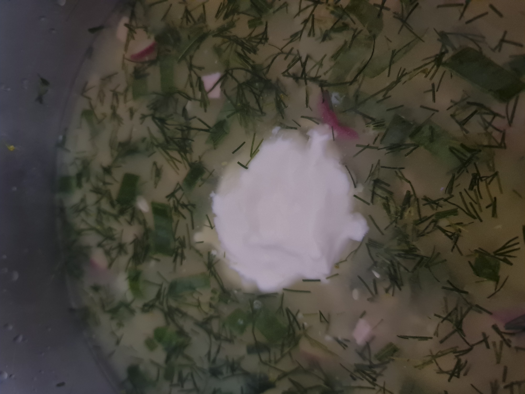The image shows a close-up, overhead view of a light-colored soup or broth in a metal pot. The soup contains chopped green herbs, likely dill, and possibly some finely diced vegetables. There are a few pieces of what appear to be radish, showing pink and white. A dollop of white sour cream or yogurt is prominently placed in the center of the soup. The environment appears to be a kitchen or dining area, but no specific location in Angarsk, Russia can be identified from the image. There are no people or discernible actions taking place, other than the presence of the prepared food.