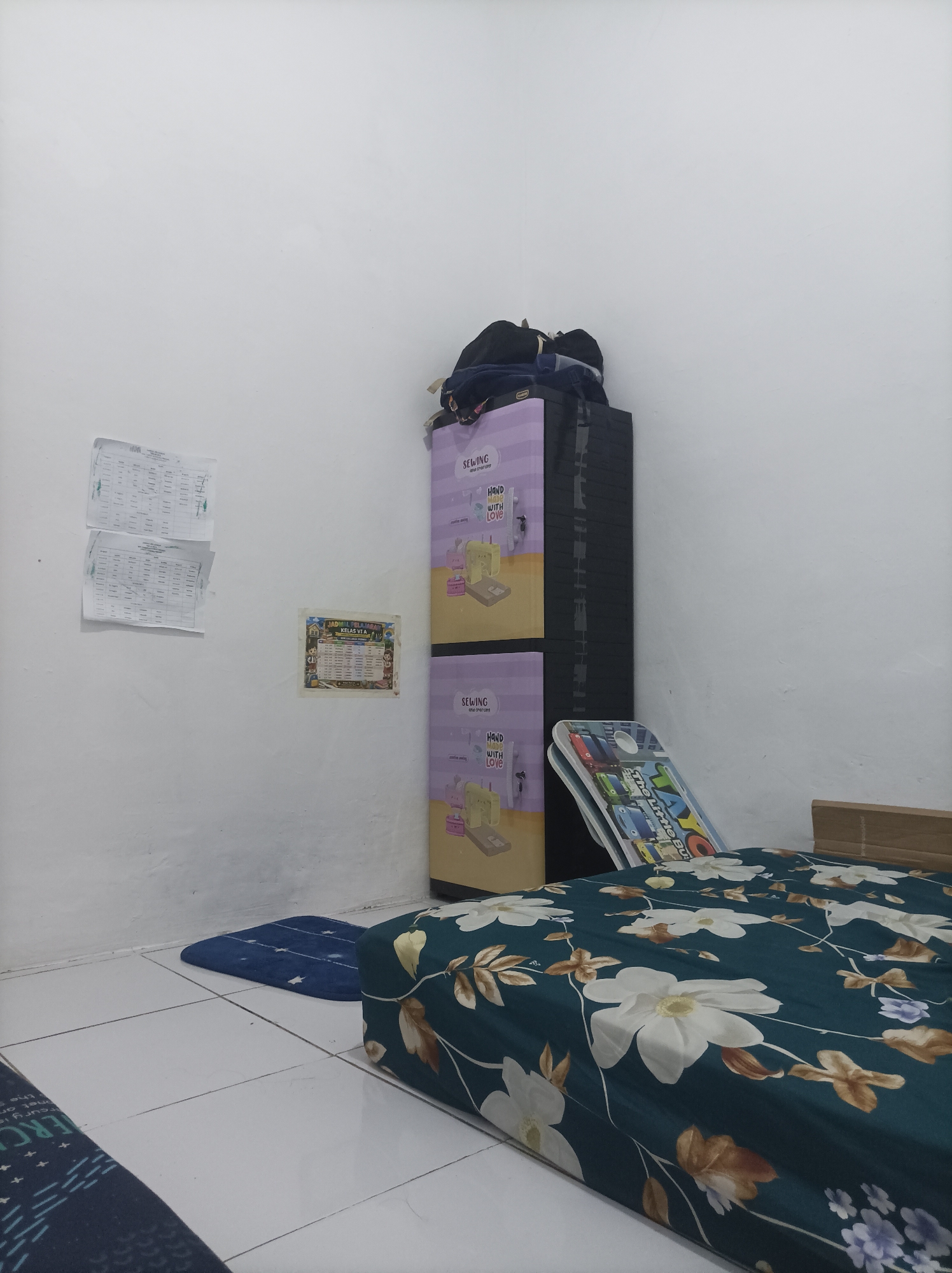 The image depicts a bedroom scene in Pamengkang, Indonesia. The room is sparsely furnished, with white tiled flooring and plain white walls. On the left wall, two schedules or timetables are pinned, and a small colorful poster is also displayed. In the center of the room, a tall, narrow, multi-tiered plastic cabinet stands against the wall, topped with some clothing items, including a black backpack. To the right of the cabinet, a children's drawing or activity board is propped up. In the foreground, a bed with a floral patterned duvet cover, featuring white and brown flowers on a dark teal background, is visible. A small blue mat with white stars is placed on the floor to the left of the bed. The lighting appears to be artificial and ambient, creating a somewhat dim atmosphere.