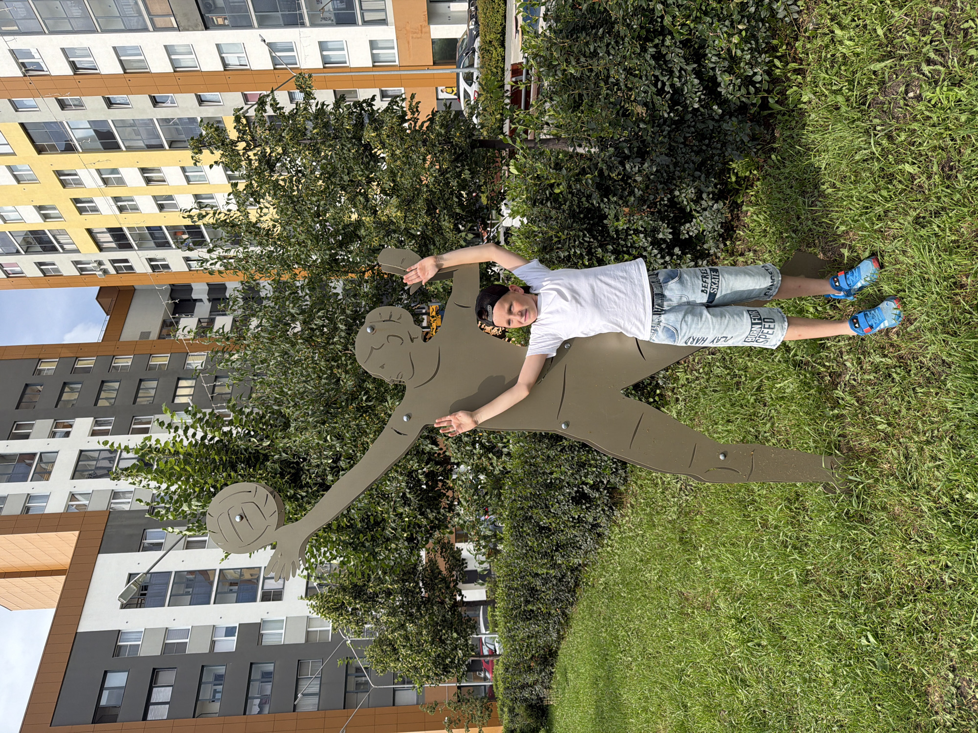 A young boy, wearing a white t-shirt, denim shorts with text "BE FREE SKATE" and "BORN FOR SPEED" printed on them, a black baseball cap, and blue sneakers, stands in front of a metal silhouette of a person playing volleyball. The silhouette is brown and appears to be a sculpture. The boy is holding his left arm up, mimicking the posture of the sculpture. He is positioned in a grassy area with lush green bushes and trees behind him. In the background, there are modern, colorful apartment buildings with balconies and windows. The sky is overcast. The scene is outdoors, likely in a residential area, possibly a park or playground within Yekaterinburg, Russia.