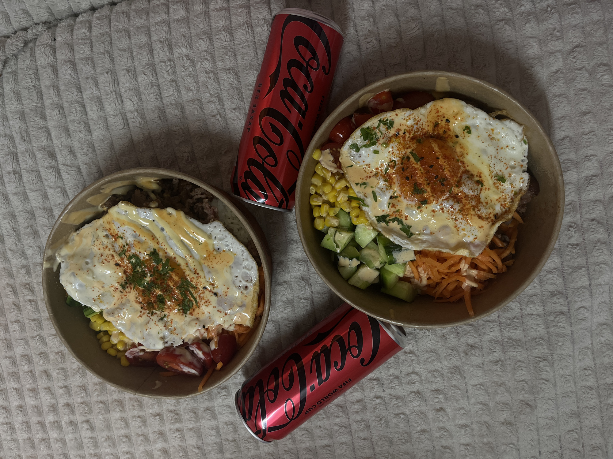 The image captures an overhead view of two similar bowls of food accompanied by two red Coca-Cola cans, all arranged on a light-colored, textured fabric surface, possibly a blanket or bedspread.
Each bowl contains a hearty mix of ingredients, topped with a fried egg. The eggs are cooked sunny-side up, featuring a firm white and a visible, slightly runny yolk. They are generously garnished with a reddish spice, possibly paprika, a sprinkle of black seeds, and fresh green herbs. A creamy, light-colored sauce is drizzled artistically over the eggs and the contents of the bowls. Beneath the eggs, various colorful ingredients are visible, including bright yellow sweet corn kernels, diced green cucumber, shredded orange carrots, and red cherry tomatoes. A darker base ingredient, likely rice or ground meat, is visible at the bottom of the bowls.
Positioned between and around the food bowls are two red Coca-Cola cans. Both cans prominently display the classic white "Coca-Cola" script logo. On the can located at the top left, Cyrillic text reading "БЕЗ САХАР" (BEZ SAKHAR), meaning "WITHOUT SUGAR" or "NO SUGAR," is visible. Additionally, both cans feature the "FIFA WORLD CUP" text horizontally, indicating a promotional tie-in.
The scene suggests a casual indoor meal, possibly a dinner or lunch, laid out in a relaxed setting. No people are visible, and the lighting indicates an indoor environment.