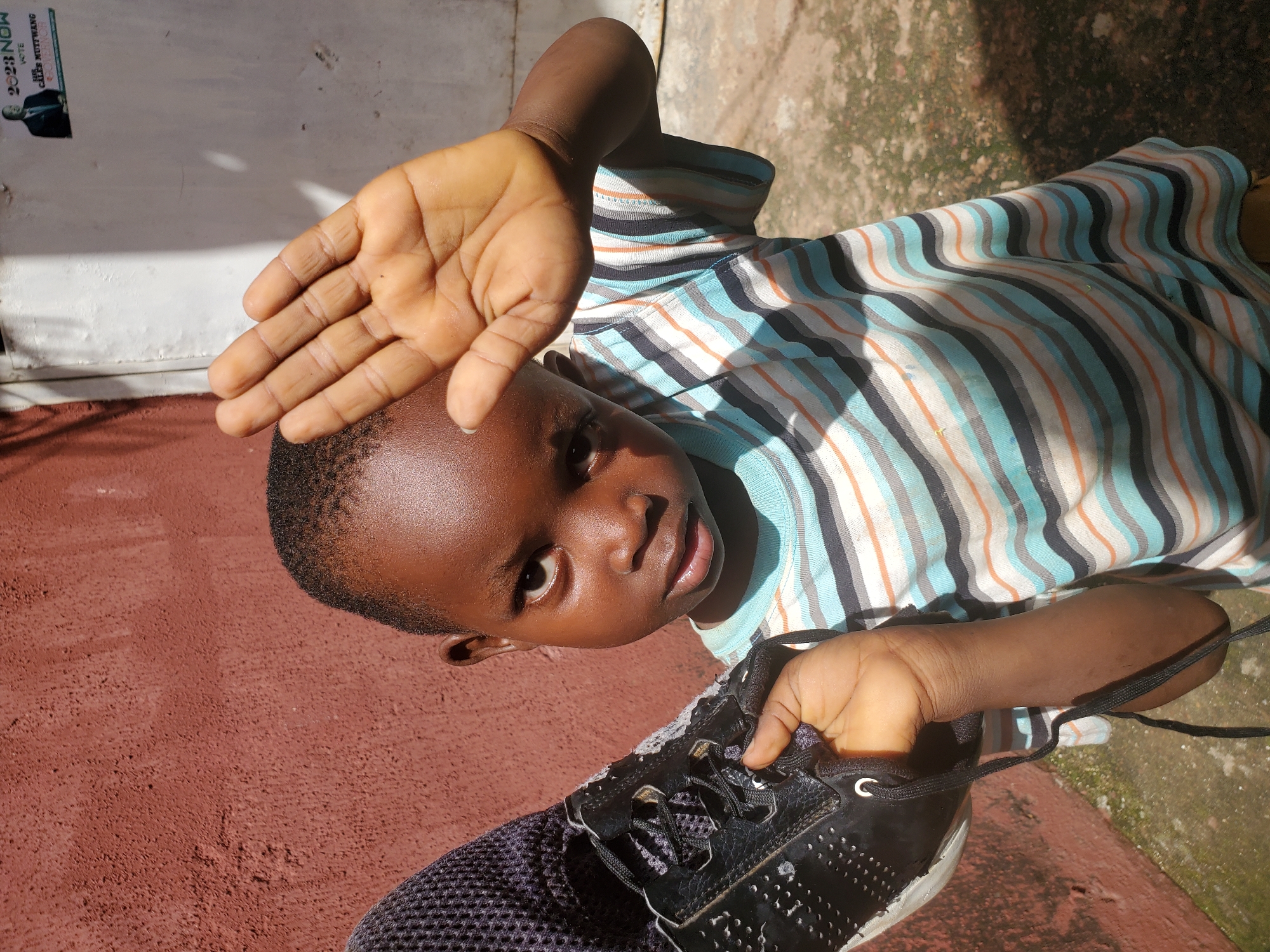 In this image, a young Black child is the central subject, positioned at an angle that emphasizes their face and a shoe held in their hand. The child is wearing a striped t-shirt with colors including blue, white, orange, and gray. Their right hand is raised, partially obscuring their face, with fingers spread open. The child's gaze is directed towards the camera, and their expression appears curious or playful. In their left hand, the child holds a black athletic shoe, with laces visible and the shoe's sole showing wear.
The background consists of a reddish-brown textured surface, likely a wall or ground, and a lighter, patchy surface above it. The lighting suggests it is daytime, with sunlight casting shadows. There's a poster or sticker visible on the lighter surface in the upper left corner of the image, featuring text that includes "2023 NOW" and a picture of a man in a suit, possibly a political campaign advertisement.
The overall scene conveys a candid and informal moment, captured outdoors. The child's interaction with the shoe and their direct look at the camera suggest a playful or engaging interaction. The location context of Jos, Nigeria, is provided, but there are no other specific visual cues to further pinpoint the location beyond general outdoor elements common in many places. The image is well-lit, with clear detail on the child and the shoe.