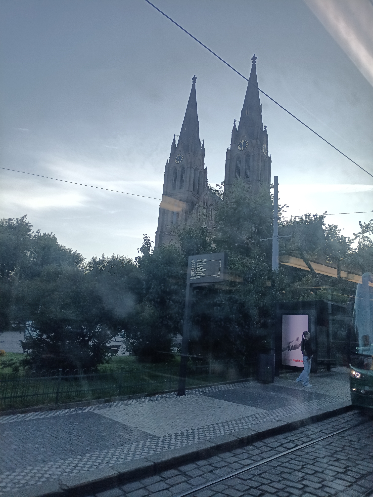 The image captures a view from inside a moving vehicle, likely a tram or bus, looking out at a street scene in Prague, Czechia. In the background, two prominent spires of a stone church rise against a cloudy sky. The church has gothic architectural elements and clocks on its towers. In the foreground, a paved walkway with a checkered pattern leads to a tram stop. A sign displaying tram routes and times is visible. To the right, a modern tram is stopped, and a person wearing jeans and a dark jacket is standing near the tram stop, looking towards an advertisement. Trees and greenery are scattered throughout the scene, and power lines are visible overhead. The overall lighting suggests it is daytime, possibly overcast.