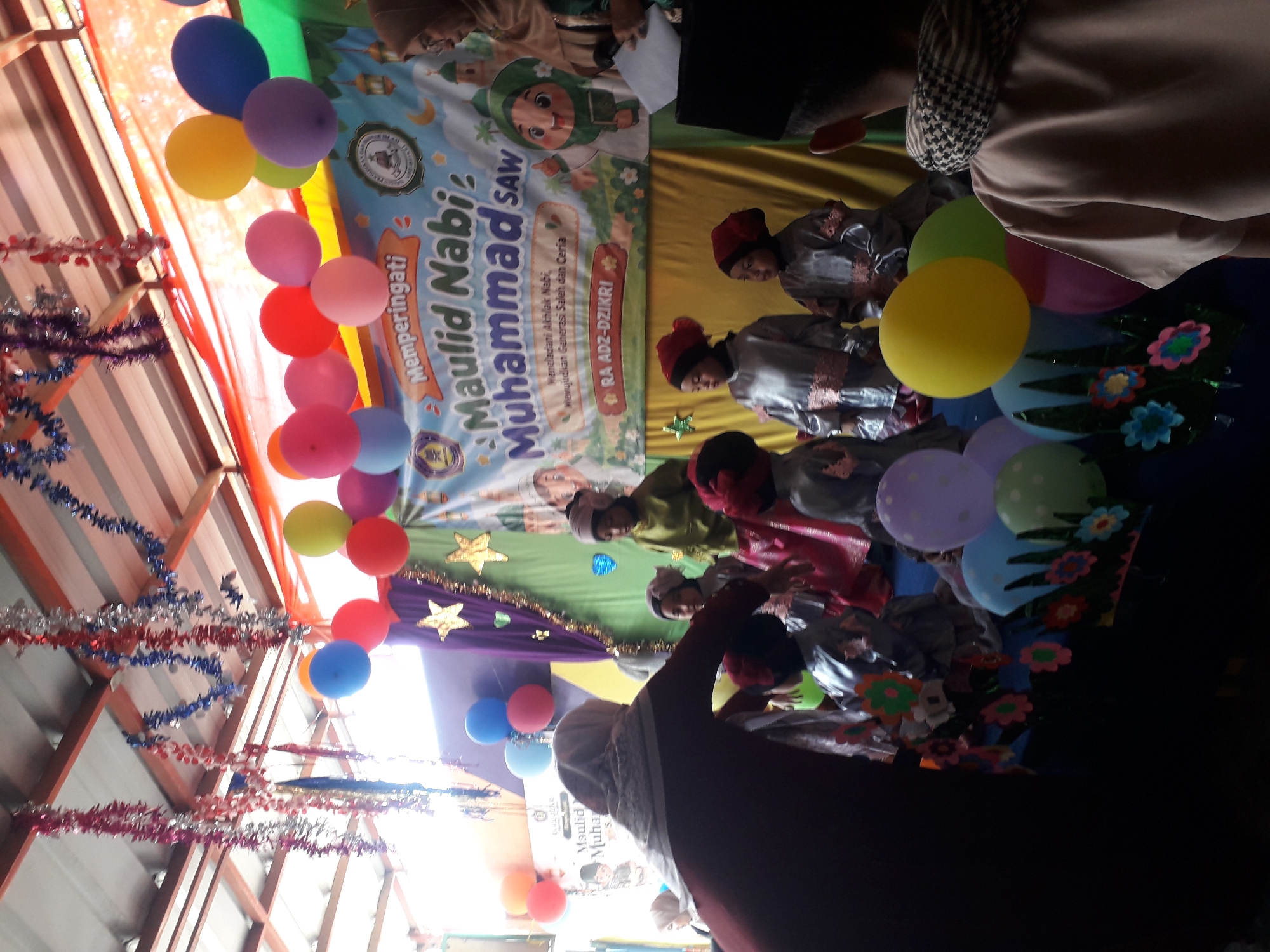 This is a horizontal, medium shot, indoor photograph taken at an event in Cankuang, Indonesia. The scene depicts a celebration for "Maulid Nabi Muhammad SAW" (The Prophet Muhammad's Birthday) at RA ADZ-DZIKRI, likely an Islamic preschool or kindergarten. The background features a banner with colorful illustrations and text related to the event. Several children, dressed in traditional attire, are gathered on a stage decorated with balloons and tinsel. Some adults are also present, including a woman in the foreground wearing a hijab, who appears to be interacting with the children. The overall atmosphere suggests a festive and communal gathering.