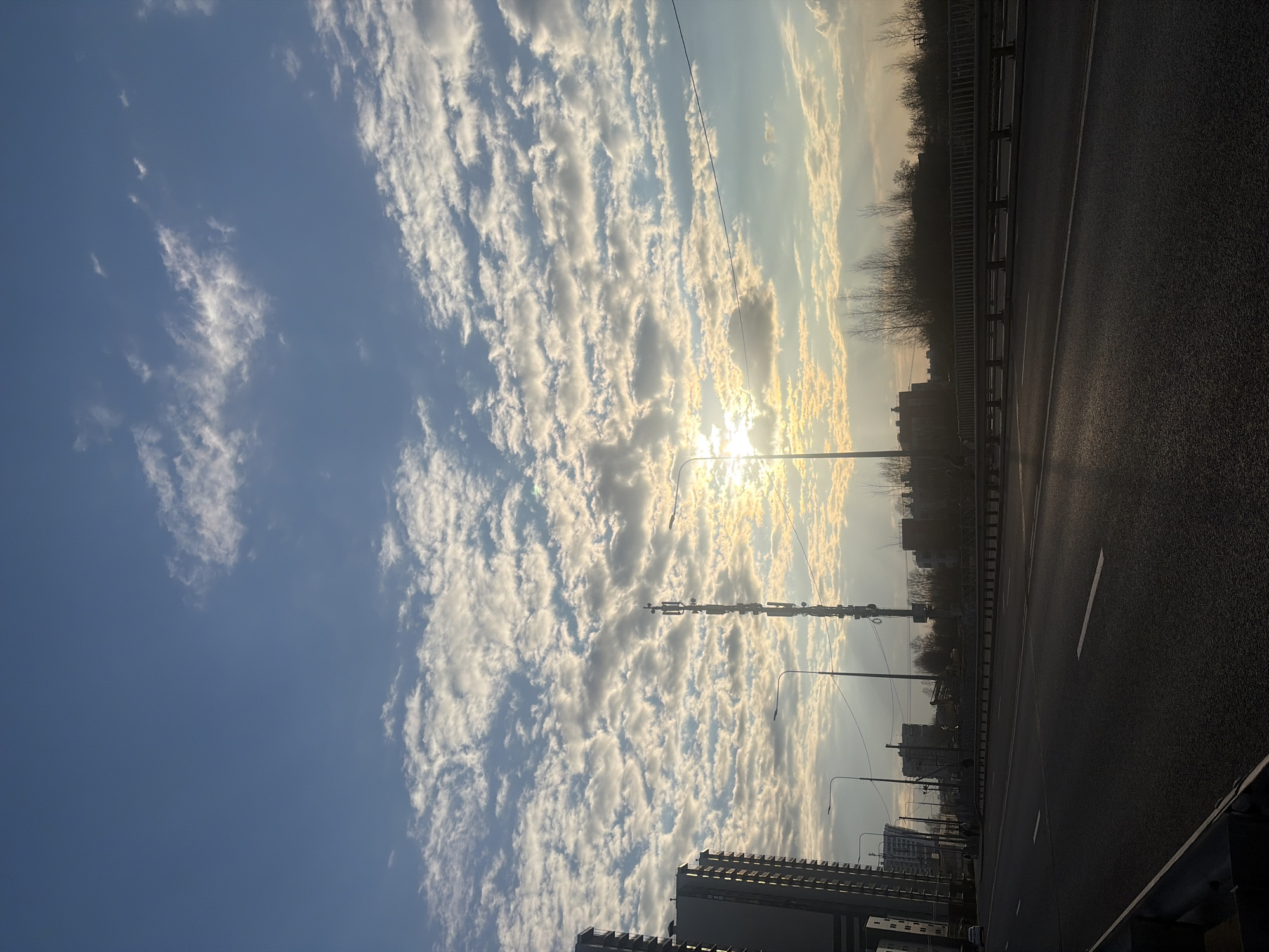 The image shows an asphalt road, possibly a highway or bridge, with a metal guardrail on the right side. Beyond the guardrail, there are trees and building silhouettes. The sky is filled with striated clouds, with the sun shining through them, creating a bright glare. There are streetlights and some buildings visible below.
Based on the appearance, it is likely an urban area within a city. The composition, with the road and clouds, might suggest a location in Saint Petersburg, Russia, considering the overall architectural style and environmental characteristics often associated with the city. However, without more specific landmarks, it is difficult to confirm the exact location.