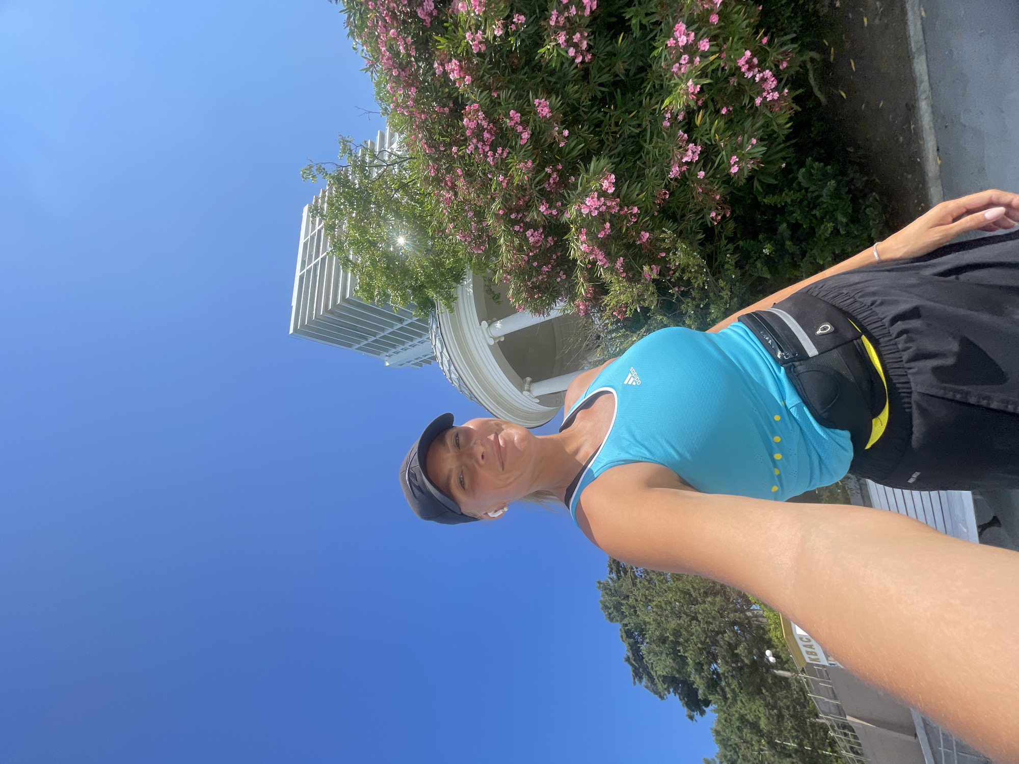 A blonde woman wearing a black baseball cap, white earbuds, a bright blue athletic tank top with yellow dots, and black athletic shorts with a black and yellow fanny pack is taking a selfie outdoors under a clear blue sky. She is smiling and looking at the camera. Behind her, to the right, is a lush green bush with numerous small pink flowers. Further in the background, to the left, is a tall, modern white building with horizontal window openings. To the left of the building and behind the woman are trees. A white circular structure with columns is partially visible behind the flower bush, and to the far left, a white bench and a yellow sign with Cyrillic lettering that appears to read "КВАСТ" are visible. The scene is likely a public park or promenade in Sochi, Russia, given the architectural styles and vegetation.