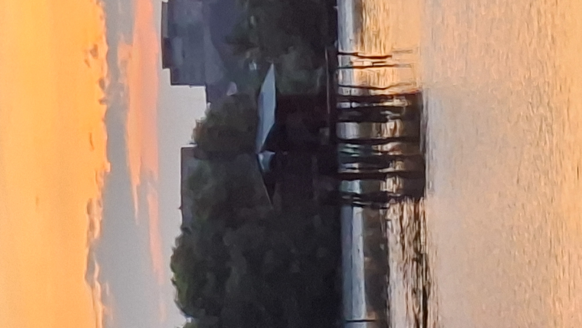 The image shows a stilt house in the water at dusk. The sky is an orange and pink gradient with wispy clouds. In the background, there are trees and buildings. The stilt house appears to be made of dark wood and has a slanted roof. The water is calm and reflects the colors of the sky. The scene is serene and tranquil. This location is likely in Lhokseumawe, Indonesia, a coastal city known for its beautiful sunsets and waters.