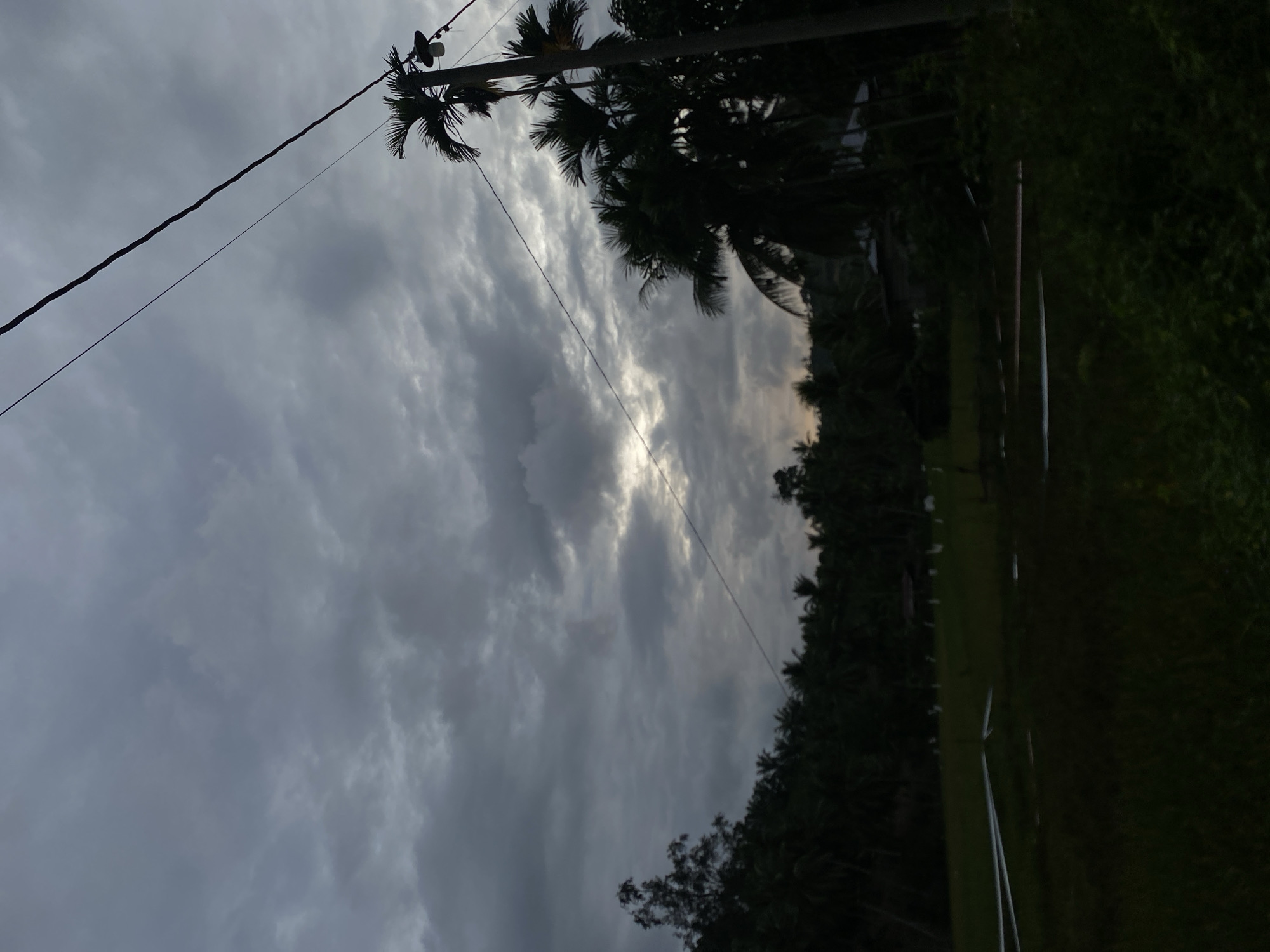 The image captures a cloudy scene in Seuneuboklhong, Indonesia. A power line pole with attached equipment is visible, supporting power lines that stretch across the frame. Palm trees and other dense vegetation line the right side of the image. The sky is filled with gray clouds, with a hint of light breaking through in the center. The lower right area is dark and indistinct.