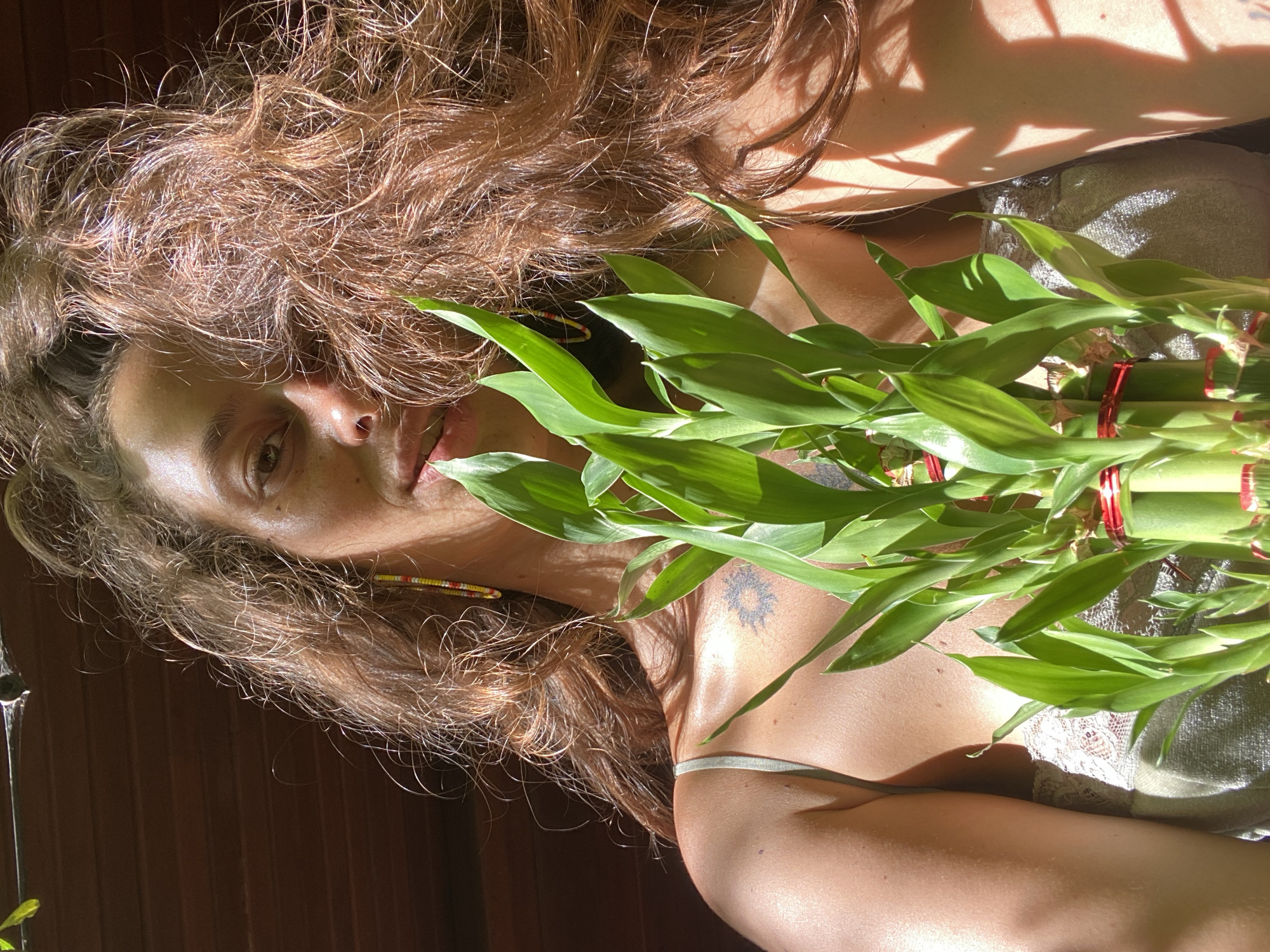 A woman is shown in a medium shot, lying down, with a bundle of bamboo shoots held in front of her. Her hair is long and wavy, with a reddish-brown hue. She has fair skin, with some freckles visible. A tattoo of a sun-like design is visible on her upper chest. She is wearing a light-colored top with lace detailing. The background appears to be a wooden surface or wall. The light source seems to be sunlight coming from above and to the side, creating shadows and highlights on her face, body, and the bamboo shoots. The scene seems to be an outdoor location in ตำบลเกาะพะงัน, Thailand.