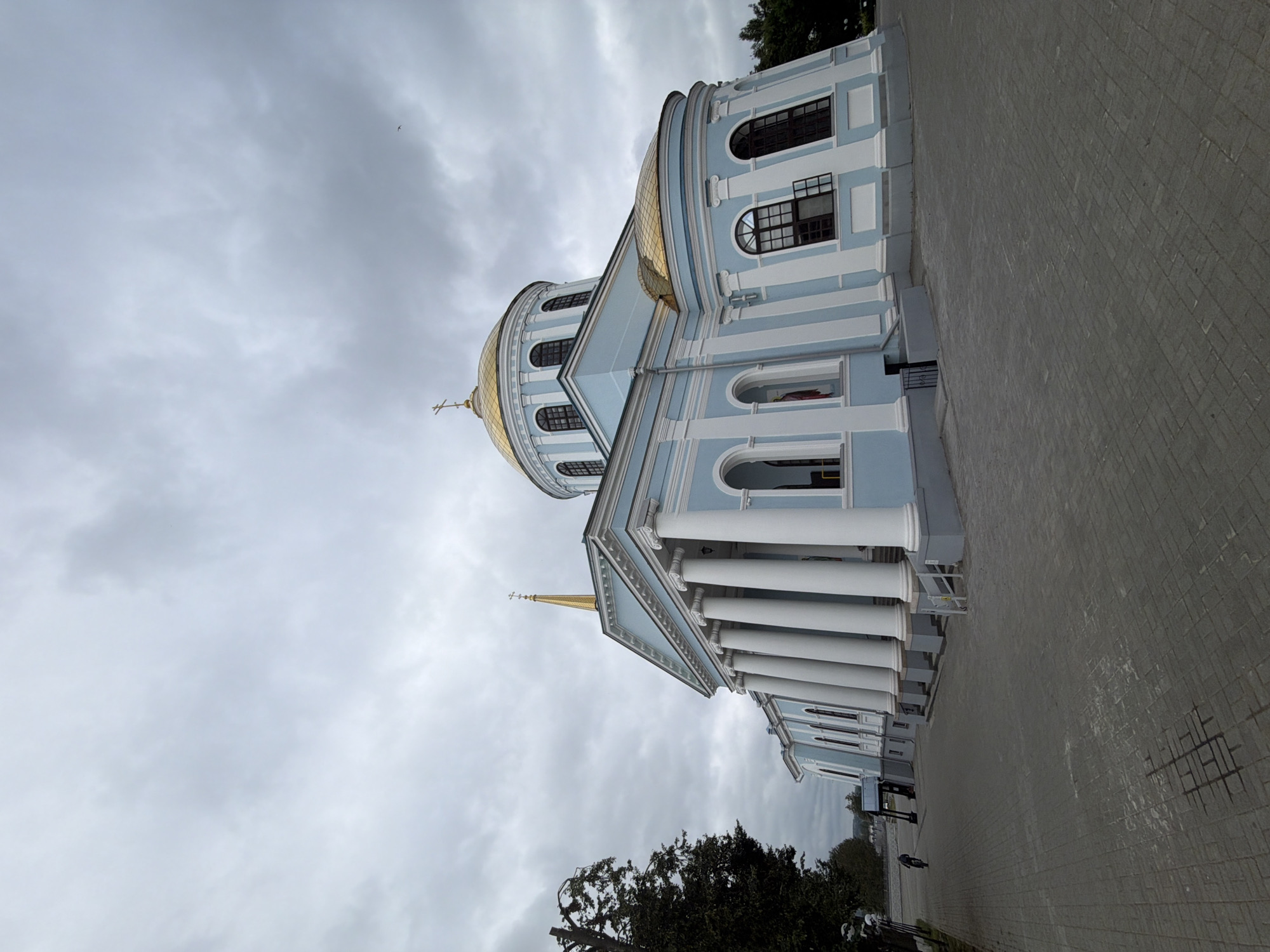 A low-angle shot captures a pale blue and white domed church with golden accents, set against a cloudy sky in Votkinsk, Russia. The church features a prominent portico with tall white columns and a triangular pediment topped with a gold spire and cross. A smaller gold dome with a cross is visible on the main structure, along with arched windows. The foreground shows a paved walkway with a person riding a bicycle in the distance. Lush green trees line the left side of the walkway. The overall atmosphere is somber due to the overcast weather.