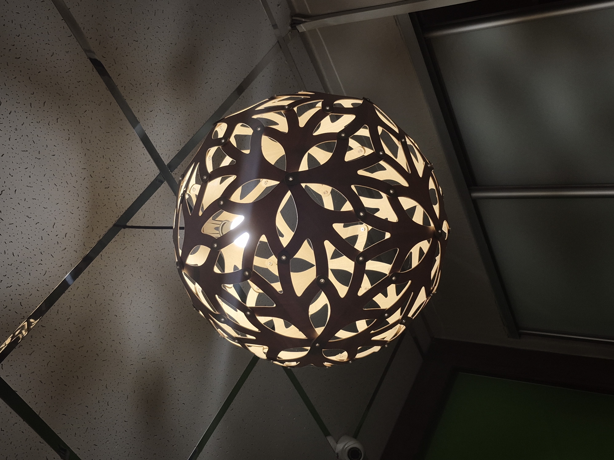 The image presents an indoor overhead view, focusing on a spherical, decorative light fixture suspended from a ceiling. The main subject, the light fixture, is crafted from a dark brown material, possibly wood or metal, featuring intricate, organic cutouts that resemble intertwined leaves or tree branches. Multiple light bulbs are visible within the sphere, emitting a bright, warm yellow glow that spills through the cutouts, casting light and shadow patterns. Small, silvery fasteners secure the components of the fixture.
The ceiling above is composed of light-colored acoustic tiles with a textured, speckled pattern. Several dark, reflective metallic beams or strips diagonally cross the ceiling, forming geometric shapes and adding a structural or decorative element.
To the right side of the frame, a flat, dark gray or black panel is visible, featuring a horizontal bar running across its width. Below this, towards the bottom right, a small, white security camera (CCTV) with a dark lens is mounted, and a glimpse of a dark green surface, likely a wall or a panel, can be seen.
The scene depicts a contemporary interior, possibly a commercial establishment like a restaurant, cafe, or public lobby, characterized by its modern design and functional ambient lighting. No people or activities are visible. The time of day is not discernible due to the artificial lighting. There is no visible text in the image. The setting is located in Chaikovsky, Russia.
