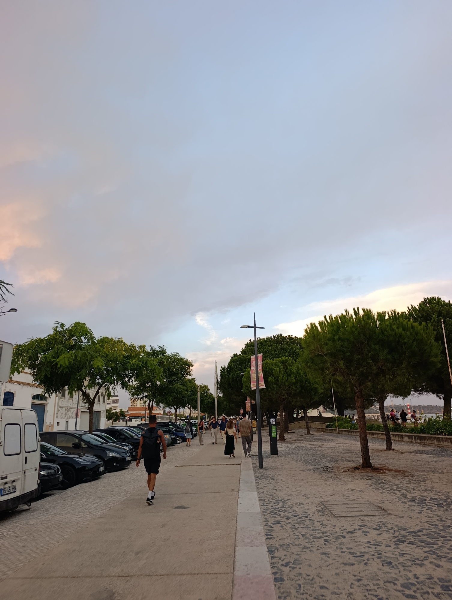 The image depicts a waterfront promenade in Setúbal, Portugal, with a view of the Sado River estuary under a cloudy sky tinged with pink and orange hues. On the left, a white van is parked alongside a row of dark-colored cars. A man in a black tank top and shorts is walking away from the camera on the paved walkway. Further down the promenade, a line of trees borders the cobblestone path, and several people are visible strolling or standing near the water. In the distance, a lighthouse is discernible. The scene suggests a late afternoon or early evening setting.