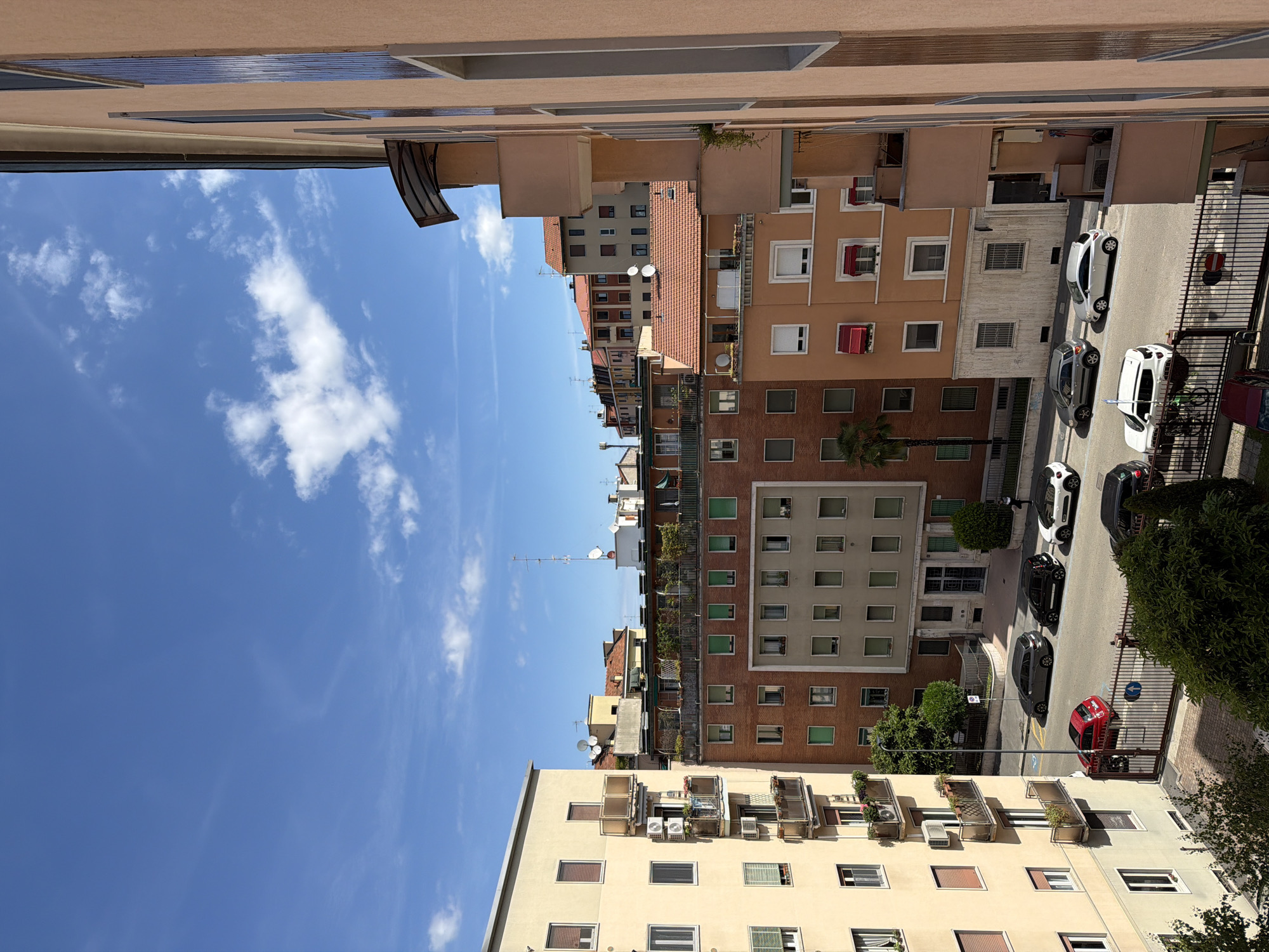 The image is an eye-level, vertical-oriented photograph of a street scene in Milan, Italy. The buildings on either side of the street are typical multi-story residential structures of varying heights and architectural styles. The building on the left is painted in pale yellow or beige tones, with windows framed in darker shades of brown or gray. Several balconies are visible, some fitted with air conditioning units.
The buildings on the right side of the street exhibit a variety of colors, from brick red to peach and beige. Some windows are adorned with awnings or shutters, and there are some hints of greenery. The street below is lined with parked cars, a mix of black, white, and red vehicles. The road is relatively narrow, with a metal fence running along the sidewalk on the right side.
Above the buildings, a blue sky with scattered white clouds is visible. There are also numerous satellite dishes, and antennas fixed atop the buildings.