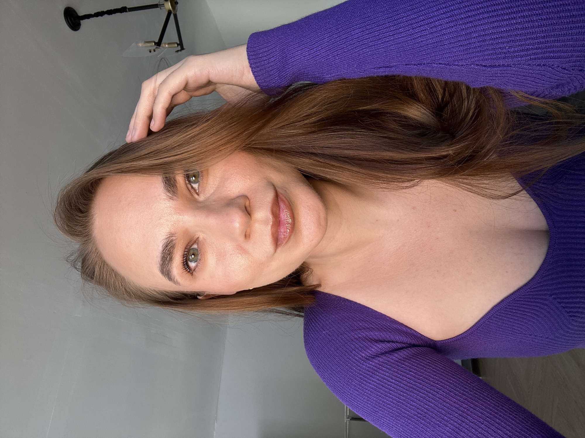A woman with brown hair and green eyes is wearing a purple ribbed shirt with a sweetheart neckline. She has her hand on her head and is smiling. The background is a light grey wall, and a black and gold light fixture is visible in the upper left corner.