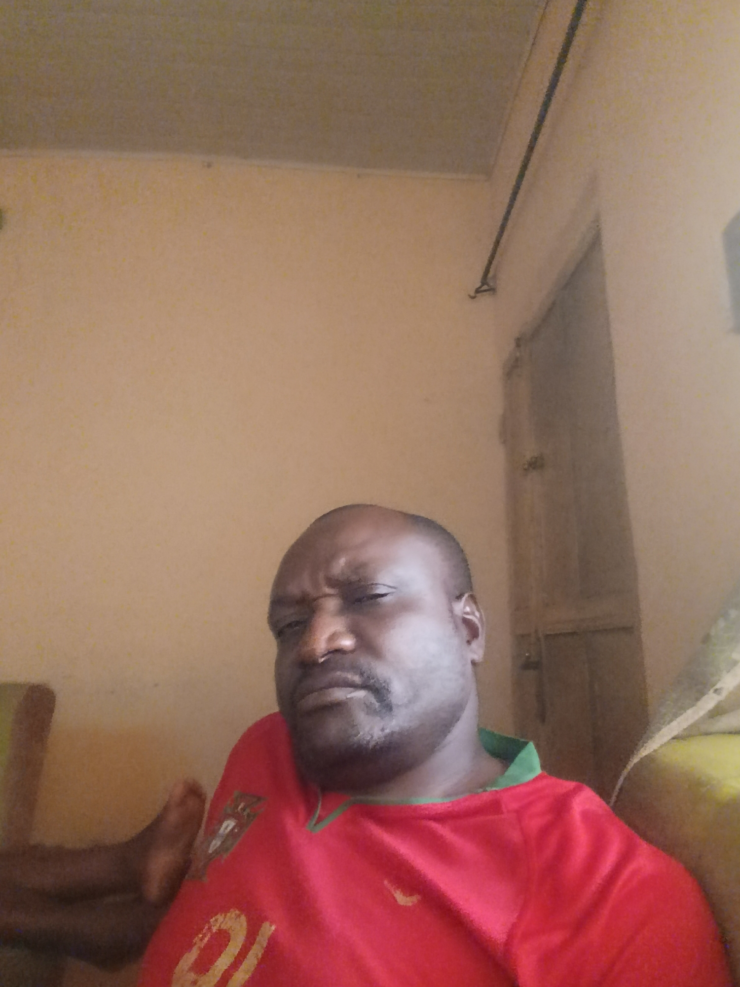 This is a medium shot, self-taken photograph of a man with dark skin and a shaved head. He is wearing a red t-shirt with a green collar and a crest on the left side of his chest, possibly a sports jersey. His expression is somewhat grimacing or showing disapproval. His left arm is visible, bent at the elbow and his hand is resting near his face. He is seated in what appears to be an indoor setting, with light beige walls and a ceiling visible in the background. To the right, a closed door with a visible handle and lock can be seen, along with what might be a closet or wardrobe. A dark horizontal rod is mounted high on the wall. To the left, part of a dark object, possibly a chair or furniture, is visible. The lighting suggests it is daytime. There is no visible text in the image, other than a faint yellow numeral that appears to be "20" or "10" on the man's shirt. The location is contextually identified as Maigizo, Nigeria.